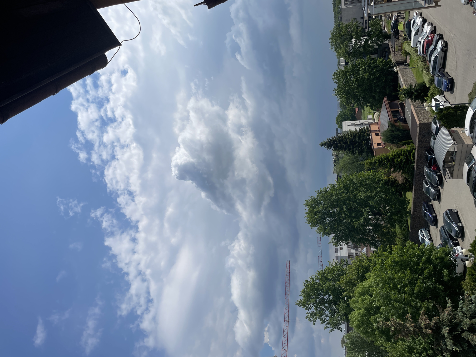 The image shows an outdoor scene in an urban area. The sky is partially cloudy with patches of blue visible between the clouds. In the foreground, the edge of a building or roof is visible. In the middle ground, there are trees and buildings. A construction crane is visible amidst the trees. A parking lot with several cars is in the background.