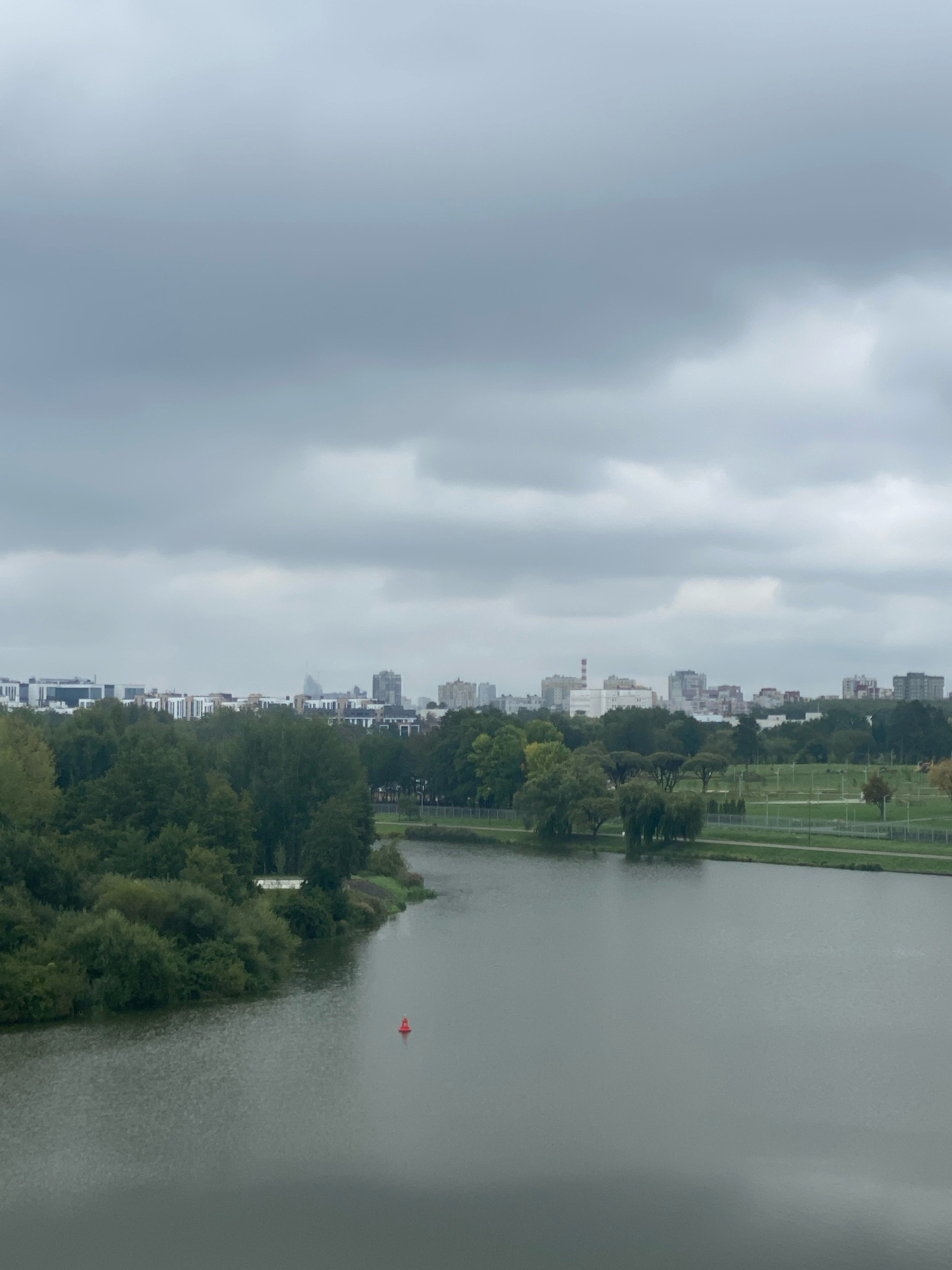 The image shows a wide shot of a body of water, likely a river or lake, in an urban setting. The water is a dull gray, reflecting the overcast sky. On the left bank of the water, there is a dense area of green trees and bushes. Further inland, a cityscape is visible with various buildings, including modern apartment complexes and some industrial structures. The sky is heavily clouded, contributing to the muted lighting of the scene. In the water, a single red buoy is visible near the left bank. The right bank of the water appears to be a park or recreational area with open green spaces and some trees. This scene is consistent with the appearance of the Svislach River in Minsk, Belarus, specifically an area where it flows through or near urban parks and residential districts. There are no people or animals visible in the frame.