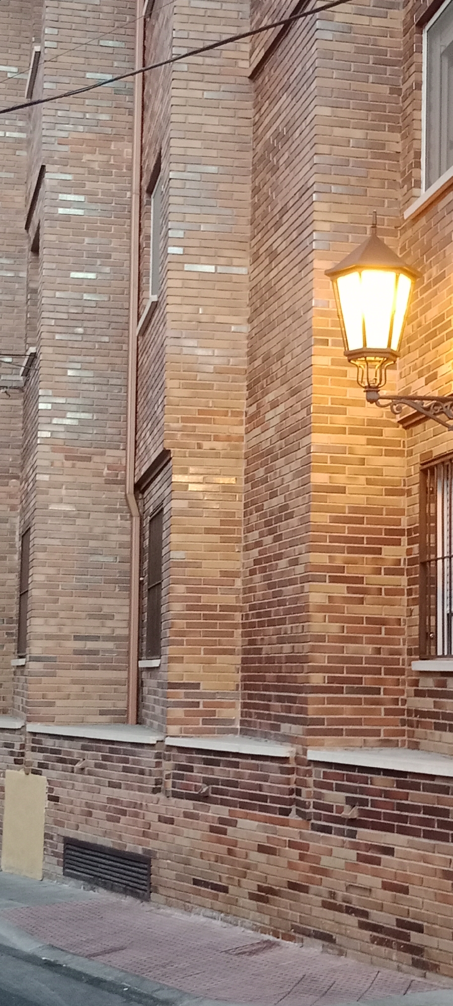 The image shows the exterior brick wall of a building with a vintage-style street lamp. The building is constructed with bricks of various shades of brown and red, arranged in a horizontal pattern. A copper downspout runs vertically along the corner of the building. There are several windows visible, some with dark frames and bars. The street lamp is illuminated and casts a warm yellow glow on the brickwork. The foreground shows a sidewalk made of reddish textured tiles and a gray asphalt road. The scene appears to be located in Galapagar, Spain, likely on a street with residential or commercial buildings. There are no people visible in the image.