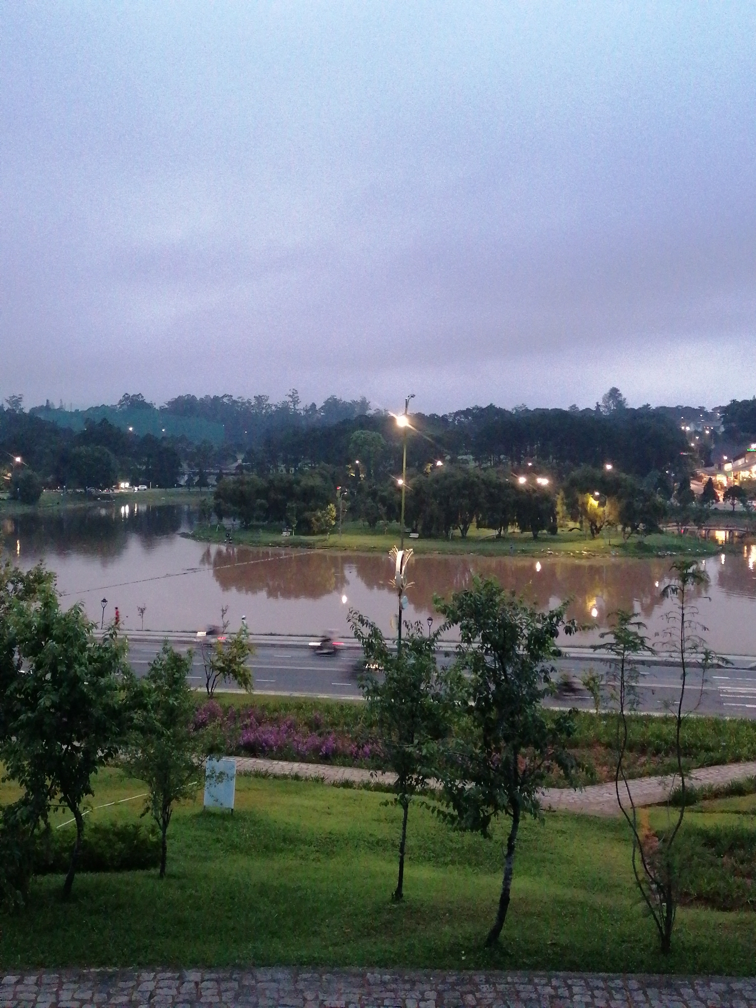 The image depicts Xuan Huong Lake in Da Lat, Vietnam, during twilight. The sky is overcast and a pale blue-gray. The lake's surface is murky brown and reflects the lights from the city, streetlights, and surrounding trees. A road runs along the edge of the lake, with several blurred vehicles suggesting movement. To the left of the road, a paved path winds through a grassy area with several trees. In the foreground, a cobblestone path is visible. The overall atmosphere is serene and somewhat moody due to the dim lighting and the subdued colors.