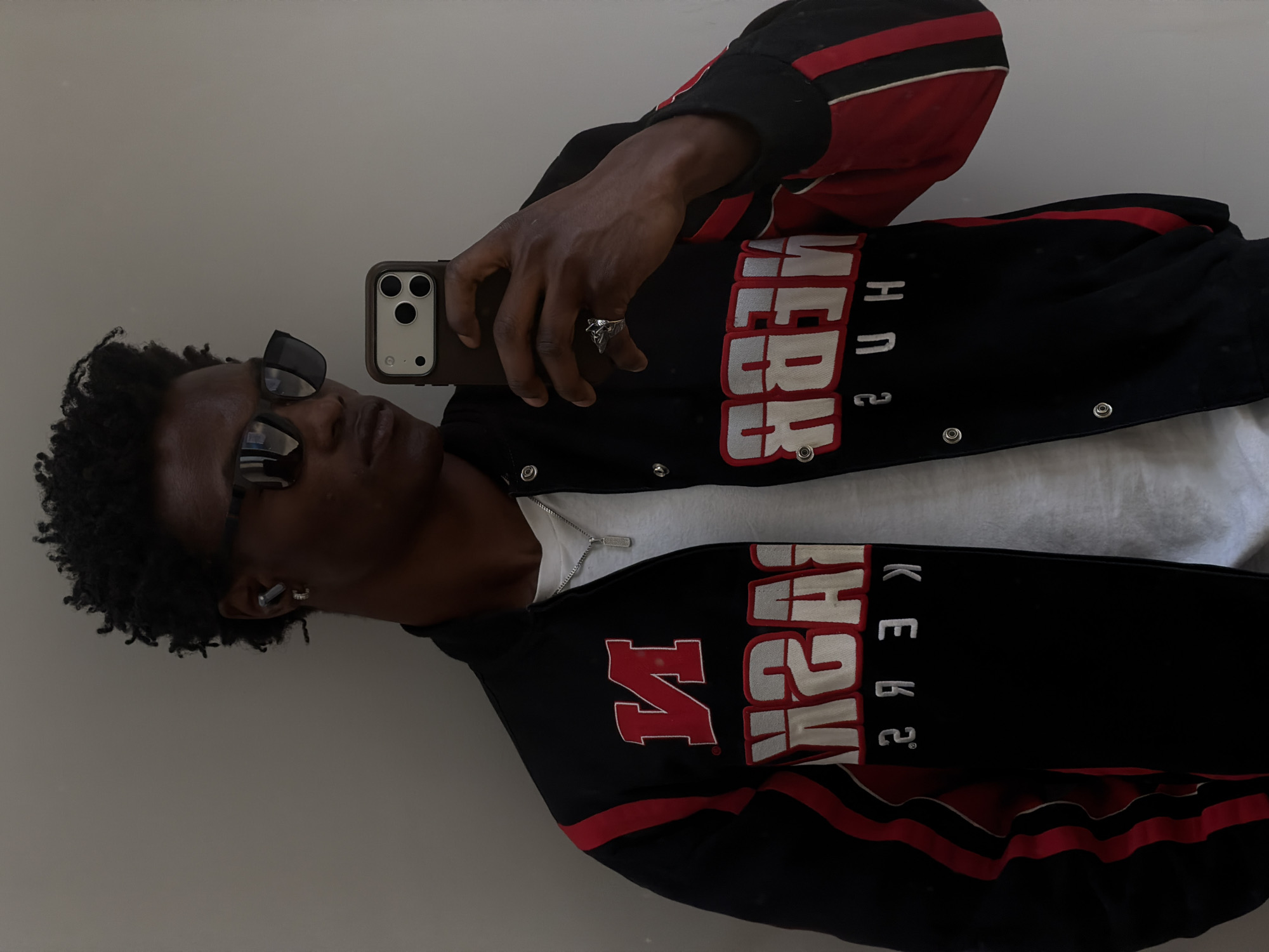 A man wearing sunglasses and a black jacket with red and white lettering is taking a selfie. He is wearing a white t-shirt underneath the jacket, and a silver ring on his finger. The background is a plain white wall. The image does not provide any information about the location in the city of London, United Kingdom.