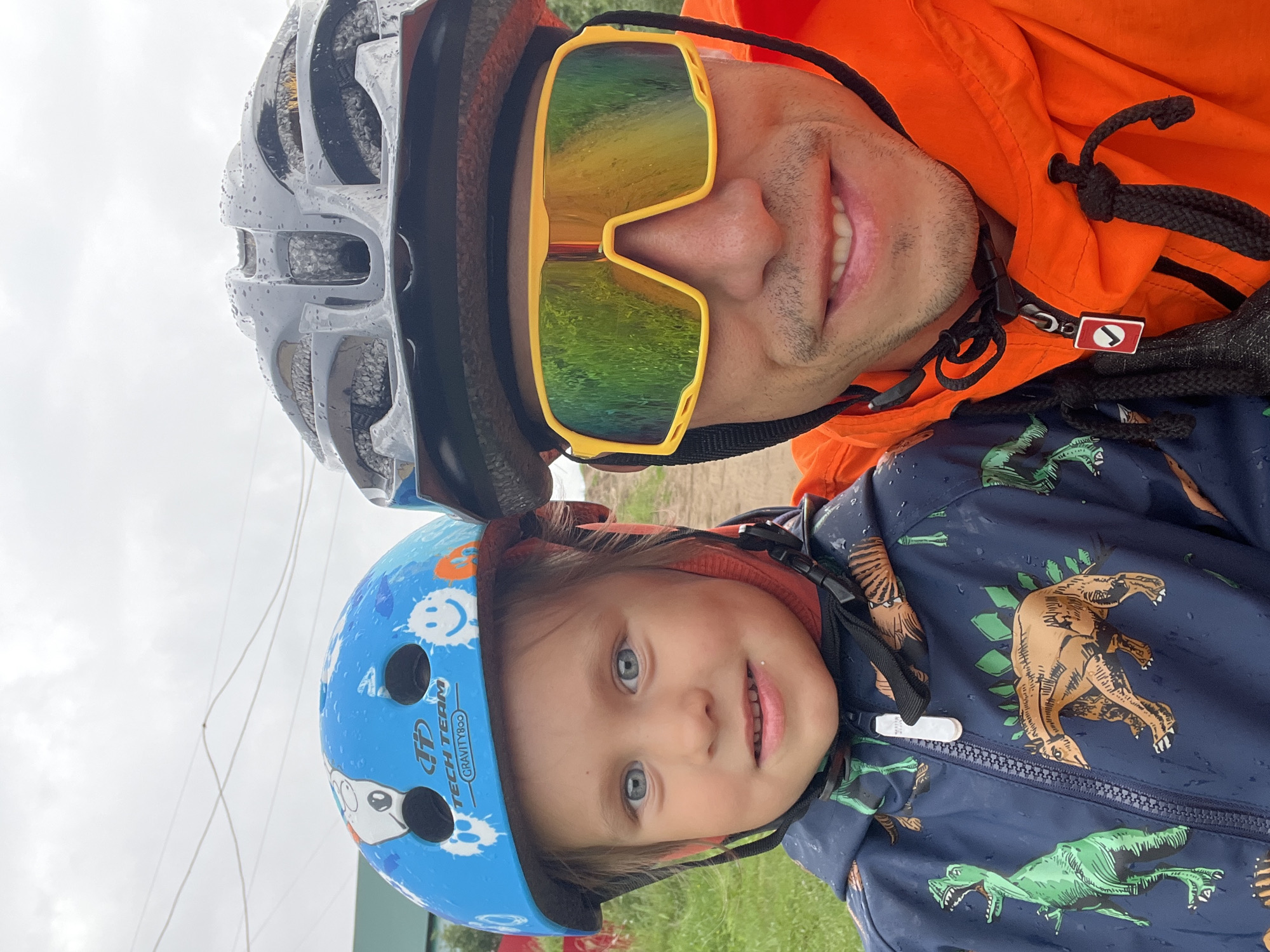 A close-up, slightly downward-angled selfie shows two individuals, an adult and a child, posing outdoors on a cloudy day. The adult, on the right, wears a black helmet and bright orange jacket, with yellow-framed, reflective sunglasses covering their eyes. They have a light beard and are smiling. The child, on the left, is wearing a blue helmet with cartoon graphics and a dark blue jacket featuring a dinosaur print. The child has blue eyes and is looking towards the camera with a slight smile. Both are equipped with protective gear, suggesting an outdoor activity such as cycling or scootering. The background is out of focus, showing a hint of greenery and overhead wires, consistent with an outdoor urban or suburban setting. The presence of raindrops on the helmets and jackets indicates recent or ongoing precipitation. The overall scene suggests a moment captured during an outing in Chaikovsky, Russia.