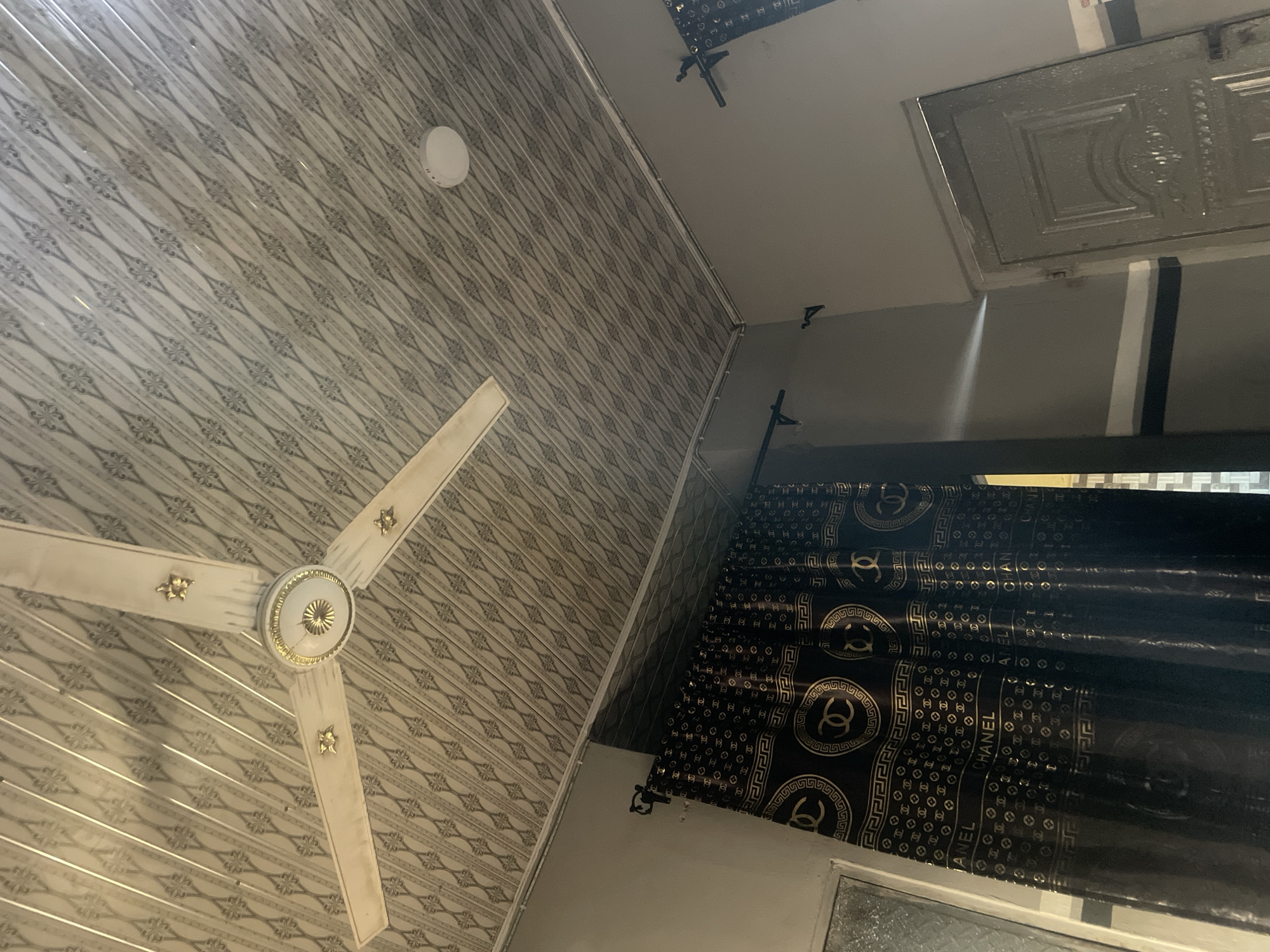In an interior shot of a room, a white ceiling fan with gold accents is visible. The ceiling is covered in a patterned material. To the right, a white circular light fixture is mounted on the ceiling. In the lower half of the image, a dark curtain with a repeating gold pattern, featuring the Chanel logo and the word "CHANEL", is hung on a black rod. To the far right, a light gray door with decorative paneling is partially visible. The walls are painted a neutral gray with a white stripe near the ceiling and a dark blue stripe below it. The scene appears to be in an indoor environment, possibly a residential or hotel room.