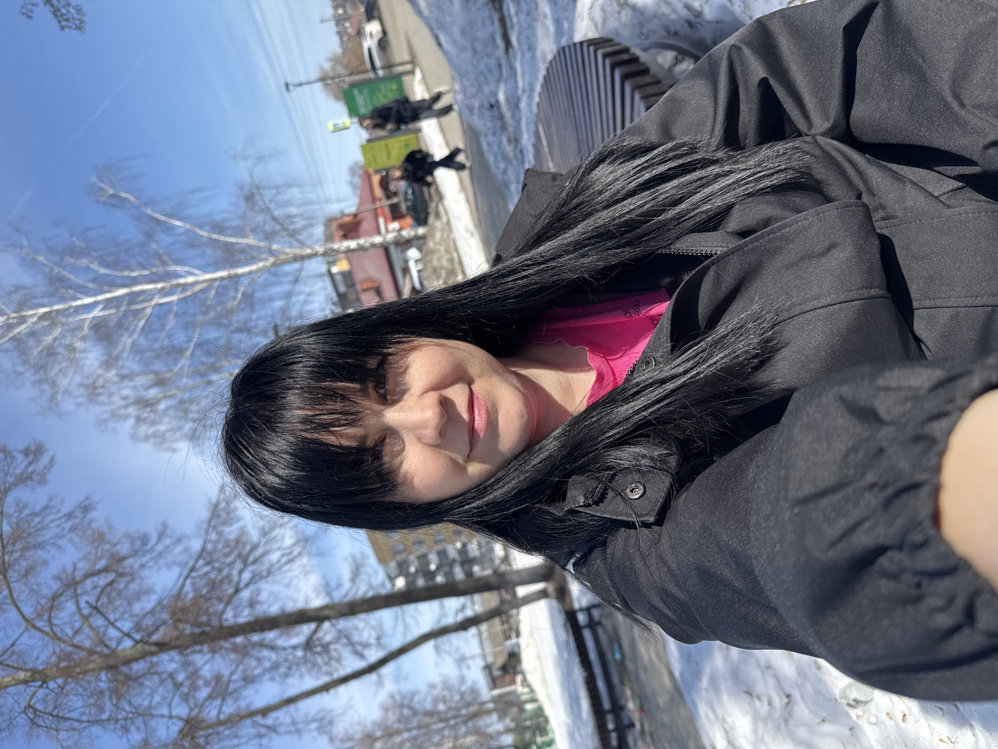 A woman with long, black hair is taking a selfie outdoors. She is wearing a black jacket and a pink shirt. The background appears to be a park or public space in winter, with patches of snow on the ground, bare trees, and urban buildings in the distance. There are figures of people walking in the background. The location is likely Zav'yalovo, Russia.
