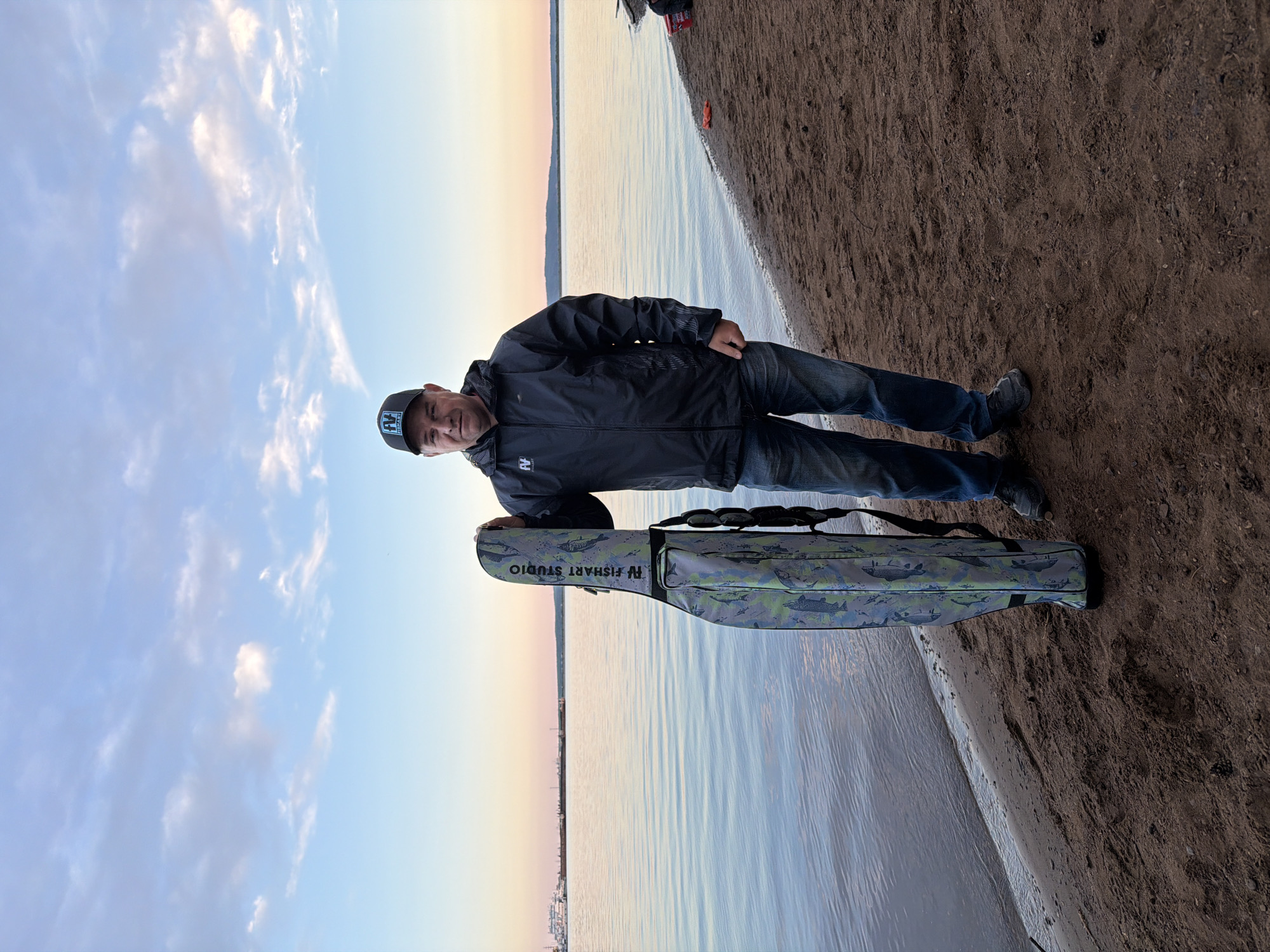 A man is standing on a sandy shoreline next to a calm body of water, likely a large river or reservoir, consistent with the location of Chaikovsky, Russia. The image captures him during the golden hour, indicated by the soft, warm light and the sky's gradient from light blue with scattered white clouds to a delicate yellowish-orange hue at the horizon. A distant landmass is visible across the water.
The man, appearing content with a slight smile, is wearing a dark jacket, blue jeans, and a dark baseball cap. The cap features a white logo and text that reads "FV FISHART". He is holding a camouflage-patterned fishing rod bag, which also prominently displays the text "FV FISHART STUDIO" and features fish illustrations as part of its design. The bag stands upright beside him, supported by his right hand.
The ground is a mixture of sand and dirt, leading to the water's edge, which shows gentle ripples. The overall scene conveys a peaceful outdoor setting, possibly at the beginning or end of a fishing trip, given the specialized equipment.