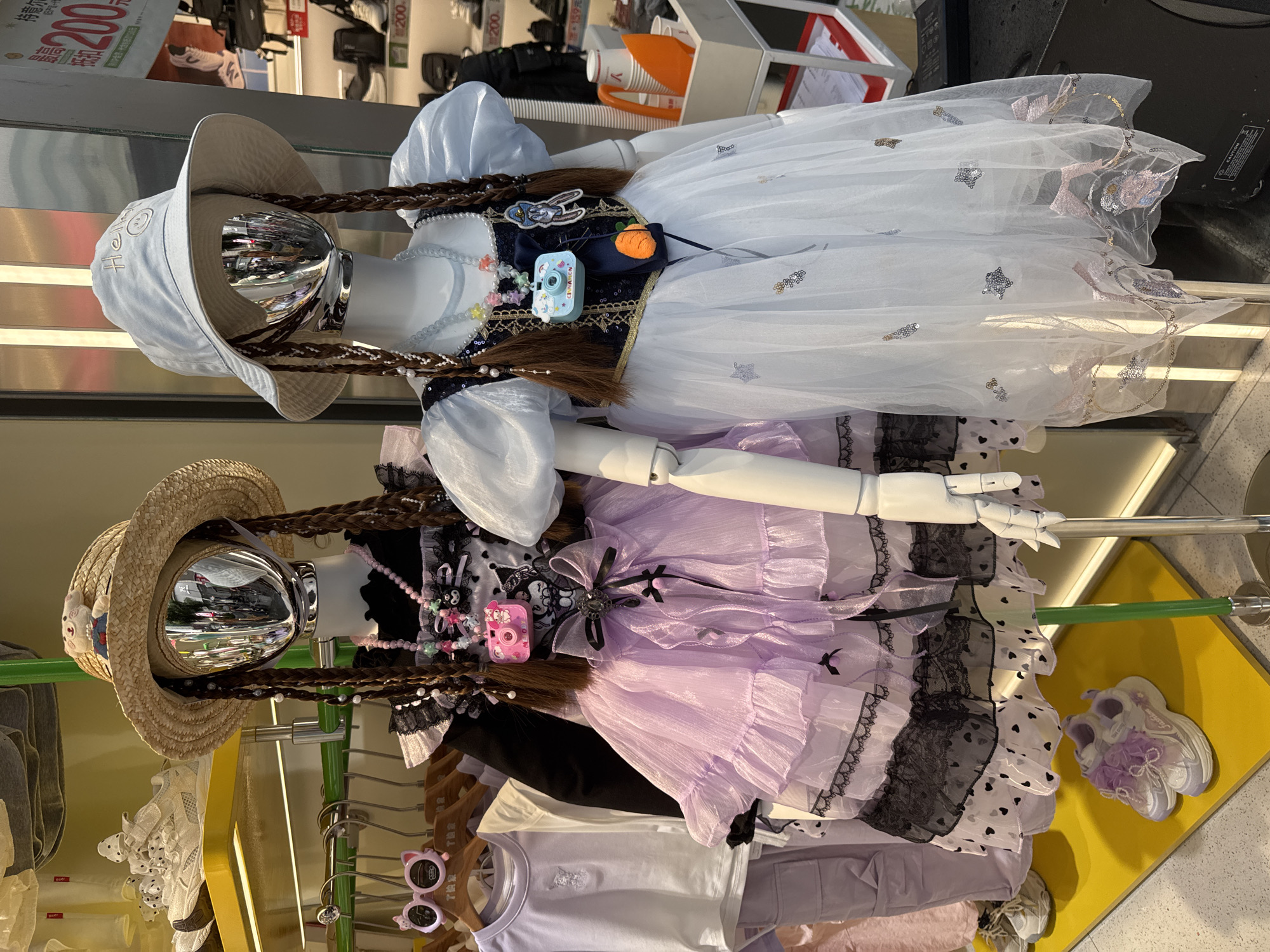 The image shows two mannequins in a store display in Jinjiang District, China. The mannequins are dressed in Lolita-style dresses with braided wigs and hats. The top mannequin is wearing a white dress with a blue bodice, embellished with a toy camera and a cartoon character. The bottom mannequin is wearing a purple dress with black lace trim. Clothes hang on a rack under the bottom mannequin. The overall environment is a retail setting, likely a clothing store.