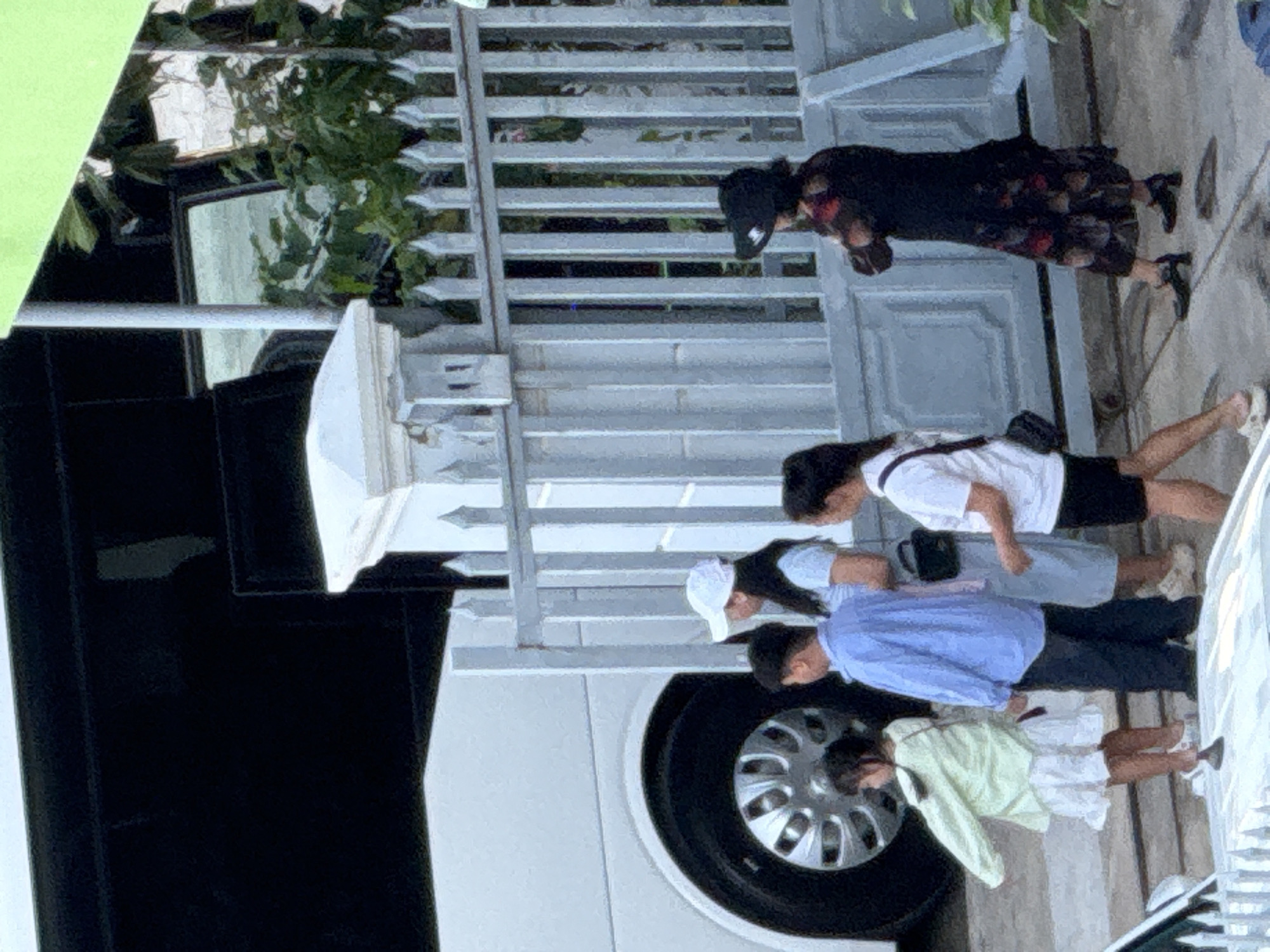 In Bac Nha Trang Ward, Vietnam, a group of individuals stands near a white bus and a decorative metal fence. A young girl in a light green long-sleeved shirt and white skirt is positioned on the left, facing away from the camera. Beside her, a boy in a blue button-up shirt and dark pants looks towards the bus. A woman wearing a white cap and a light blue dress stands behind the boy. Further to the right, another woman in a white t-shirt and black shorts is visible. A woman in a dark, patterned dress and a black hat stands near the fence, facing left. The environment includes lush green foliage behind the fence and a portion of a white bus on the left side of the frame.