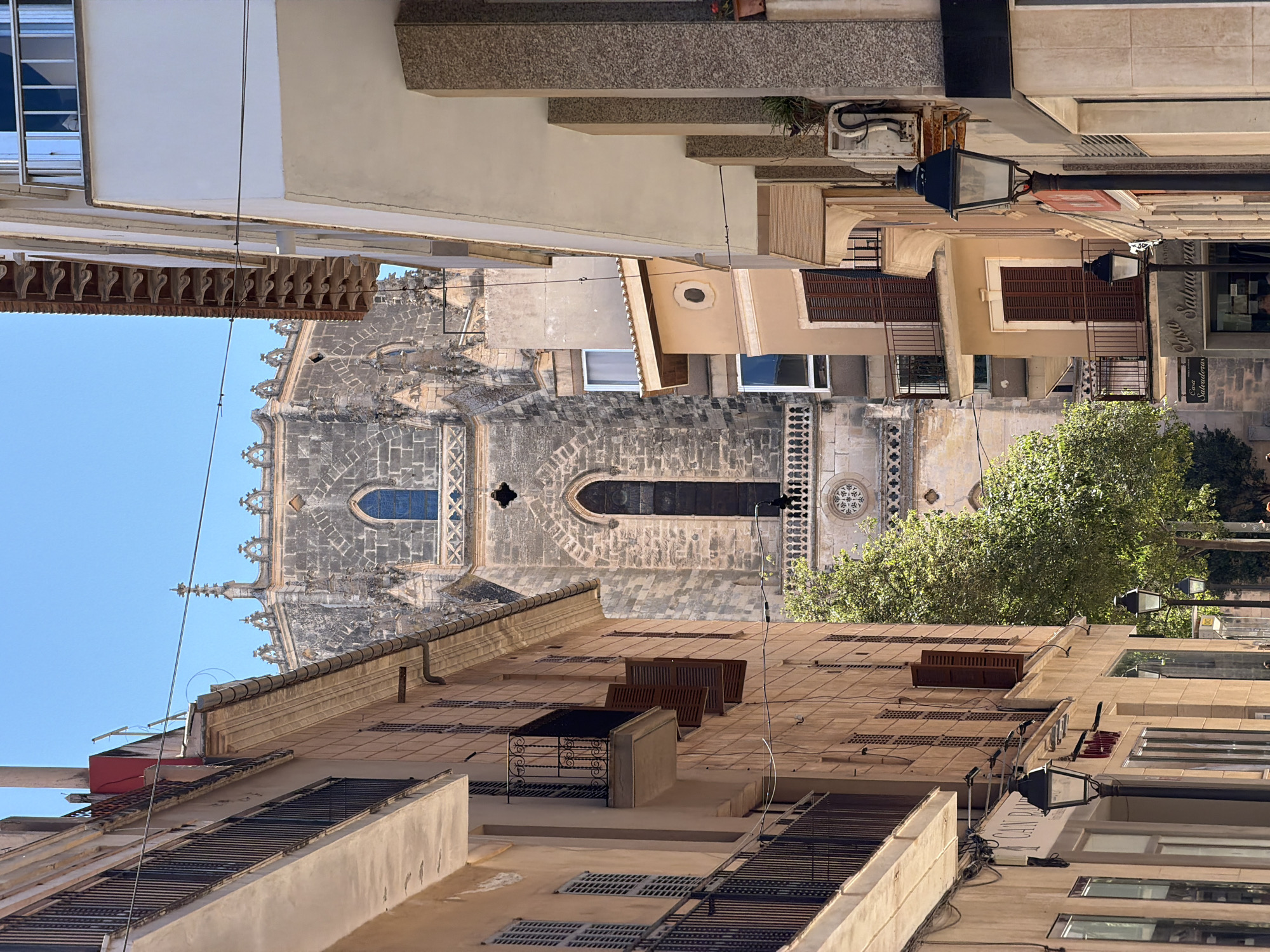 The image captures a street view in Manacor, Spain, looking towards the iconic Church of Nostra Senyora dels Dolors. The perspective is framed by the facades of residential and commercial buildings on either side of a narrow street. Sunlight illuminates the scene, casting distinct shadows and highlighting the architectural details of both the buildings and the distant church.
On the left, a building with a pale, textured facade features balconies with dark metal railings. The windows are mostly covered by wooden shutters, some of which are open, revealing glimpses of the interiors. The building has several floors, and the upper sections are partially obscured by awnings and the edge of another building.
The right side of the street is dominated by a building with a white facade, punctuated by balconies with metal railings. The ground floor appears to house commercial establishments, with visible signage and street lamps. One prominent black street lamp stands closer to the foreground, its ornate design adding to the street's character.
Towering in the background, and partially visible between the buildings, is the Church of Nostra Senyora dels Dolors. Its Gothic architecture is evident with its stone construction, arched windows, and decorative spires. The church's facade is a focal point, its intricate stonework and imposing presence contrasting with the more mundane structures of the street. A large, mature tree with dense green foliage stands in the mid-ground, partially obscuring the lower part of the church and adding a natural element to the urban landscape.
The sky above is a clear, bright blue, indicating a sunny day. The overall impression is one of a typical Mediterranean street scene, where historical architecture coexists with modern life. There are no discernible people or explicit actions taking place, creating a sense of quiet observation.