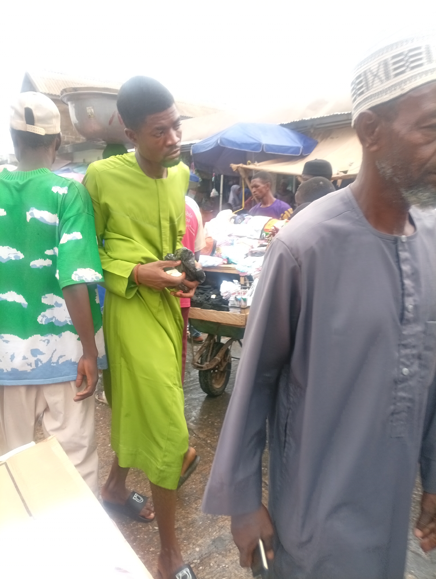 The image depicts a busy outdoor market scene in Maiburiji, Nigeria. Four individuals are prominently featured. On the left, a person wears a green t-shirt with a white cloud design and light-colored pants. Next to them, a man in a green robe holds a dark object in his hands. To the right, an elderly man wears a white kufi cap and a grey robe, holding a cell phone in his left hand. In the background, additional people are present, including a person in a purple shirt standing near a cart with items on display. An open-air structure is visible overhead, providing some shelter. The ground appears wet.