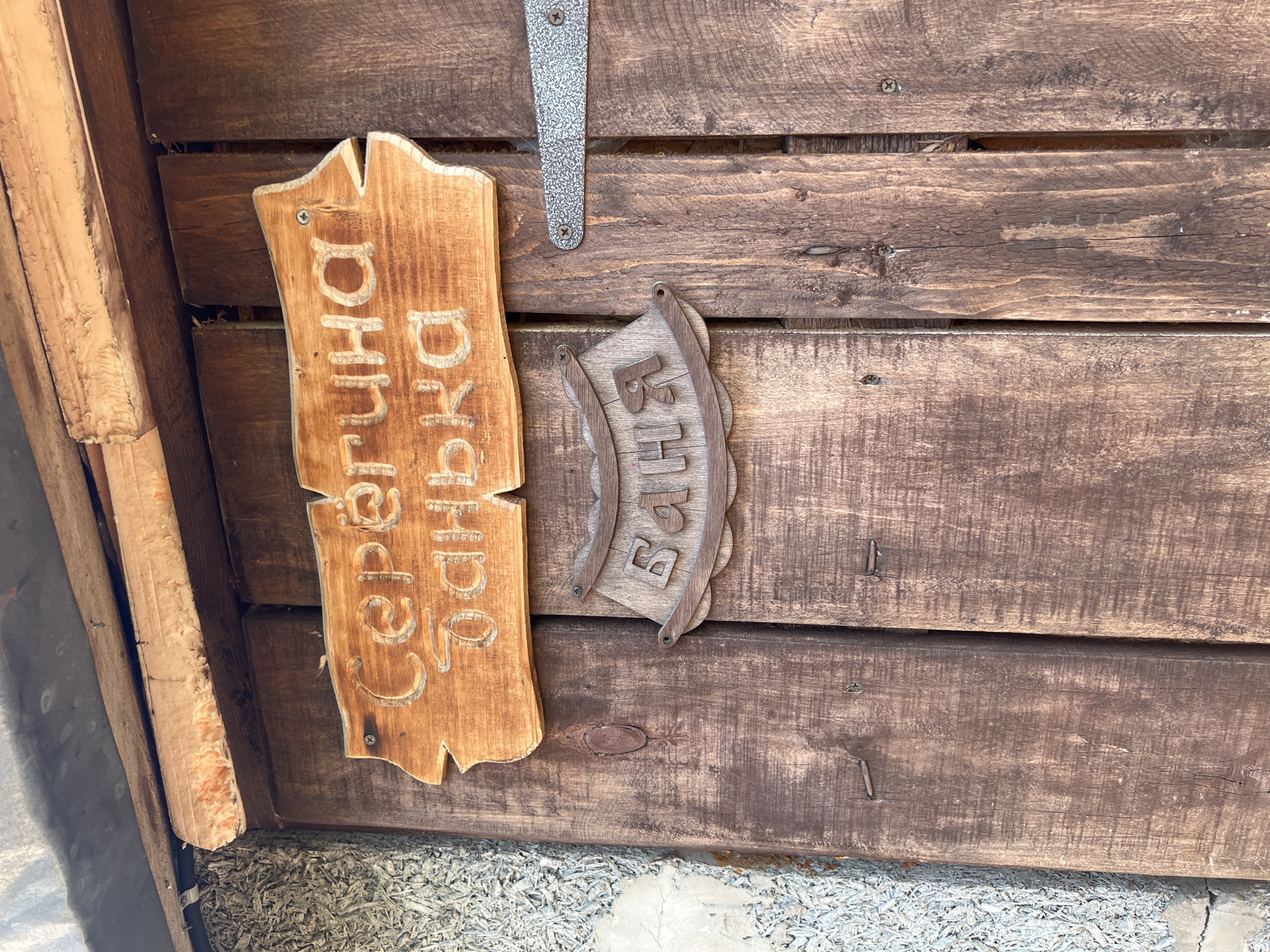 The image shows two wooden signs mounted on a wooden wall. The sign on the left has a vertical orientation and the inscription "Серегина банька" in Cyrillic script carved into it. The sign on the right is smaller, more stylized, and also has a word in Cyrillic script carved into it, possibly "Баня." A metal hinge is visible above the signs. The wall appears to be made of rough-hewn wooden planks. Based on the text on the signs, the location is likely Chaikovsky, Russia.