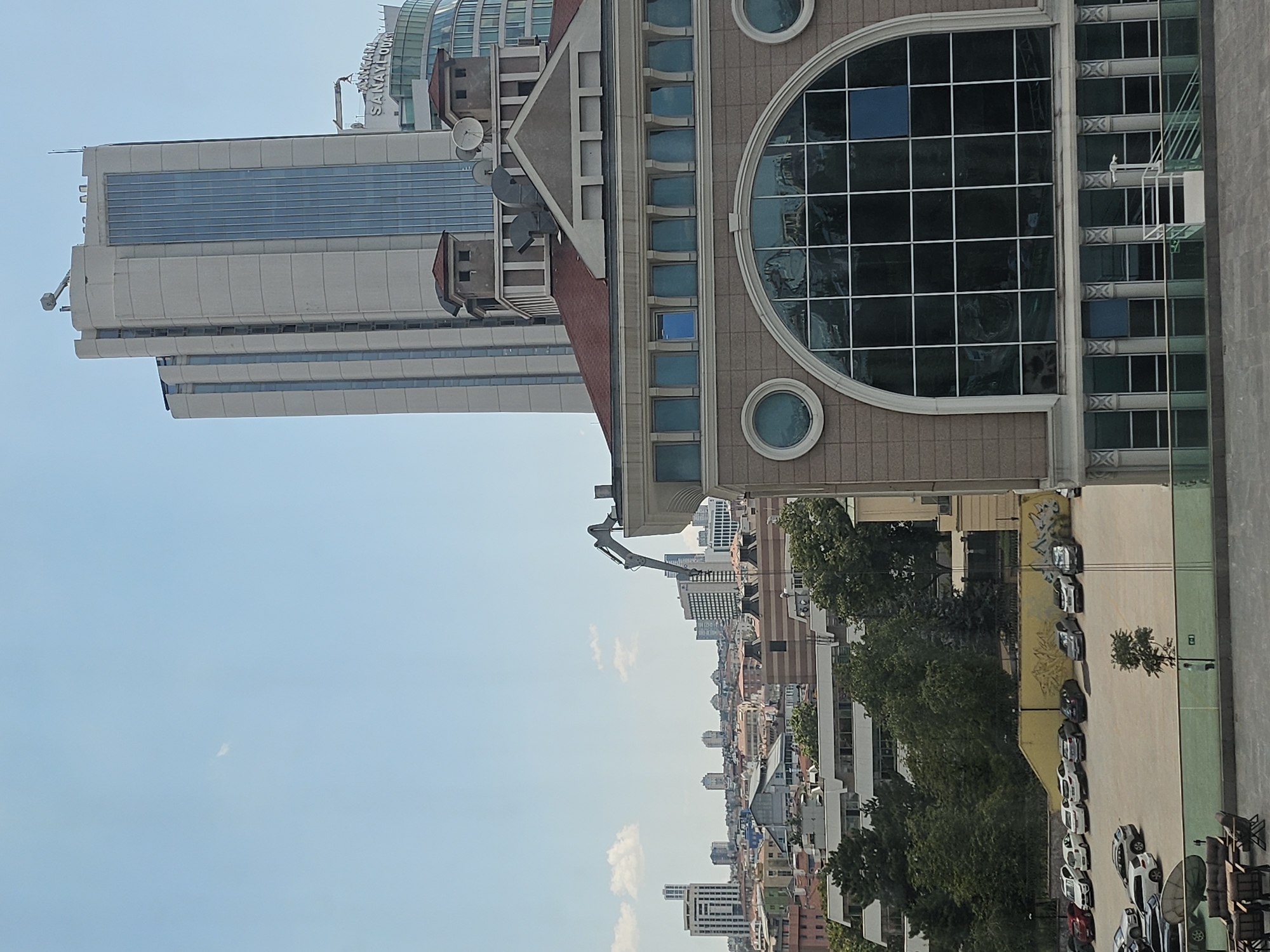 In an outdoor urban setting, the image presents a cityscape with several buildings of varying architectural styles and heights. A prominent modern skyscraper with a light-colored facade and blue vertical window panels dominates the upper right portion of the frame. To its left, another tall, light-colored building with segmented sections and some vertical windows is visible. In the foreground, a building with a red tiled roof and beige stonework features a large arched window with a grid of dark panes. Adjacent to this building, a parking lot filled with various cars, including sedans and SUVs, is visible. A section of green space with trees and a single standing person is situated beyond the parking lot, separated by a glass barrier. The sky above is a clear light blue with a few scattered white clouds. A construction crane with an extended arm is partially visible against the sky, positioned between the two prominent skyscrapers.