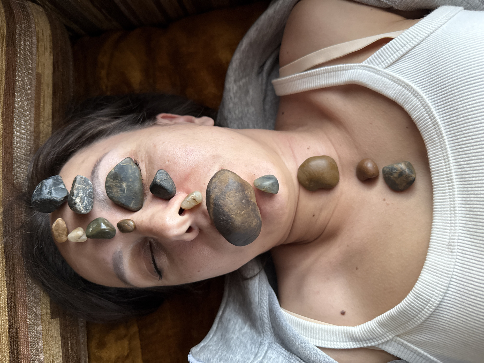 In a close-up, top-down view, a person with their eyes closed lies on a brown, textured surface, possibly a couch. Their head is tilted slightly upwards, and a series of smooth, rounded stones are meticulously arranged on their forehead, nose, and chest. The stones vary in color, from dark grey and black to lighter brown and tan, with some exhibiting interesting patterns. The person is wearing a white ribbed tank top and a grey hooded sweatshirt is draped over their left shoulder. The environment appears to be indoors, possibly a residential setting, with the background showing the textured material of the couch and a hint of a darker, out-of-focus area. There is no indication of the city of Solnechnyi, Russia, or any specific urban features in the image. The scene is static and suggests a moment of relaxation or a therapeutic practice.
