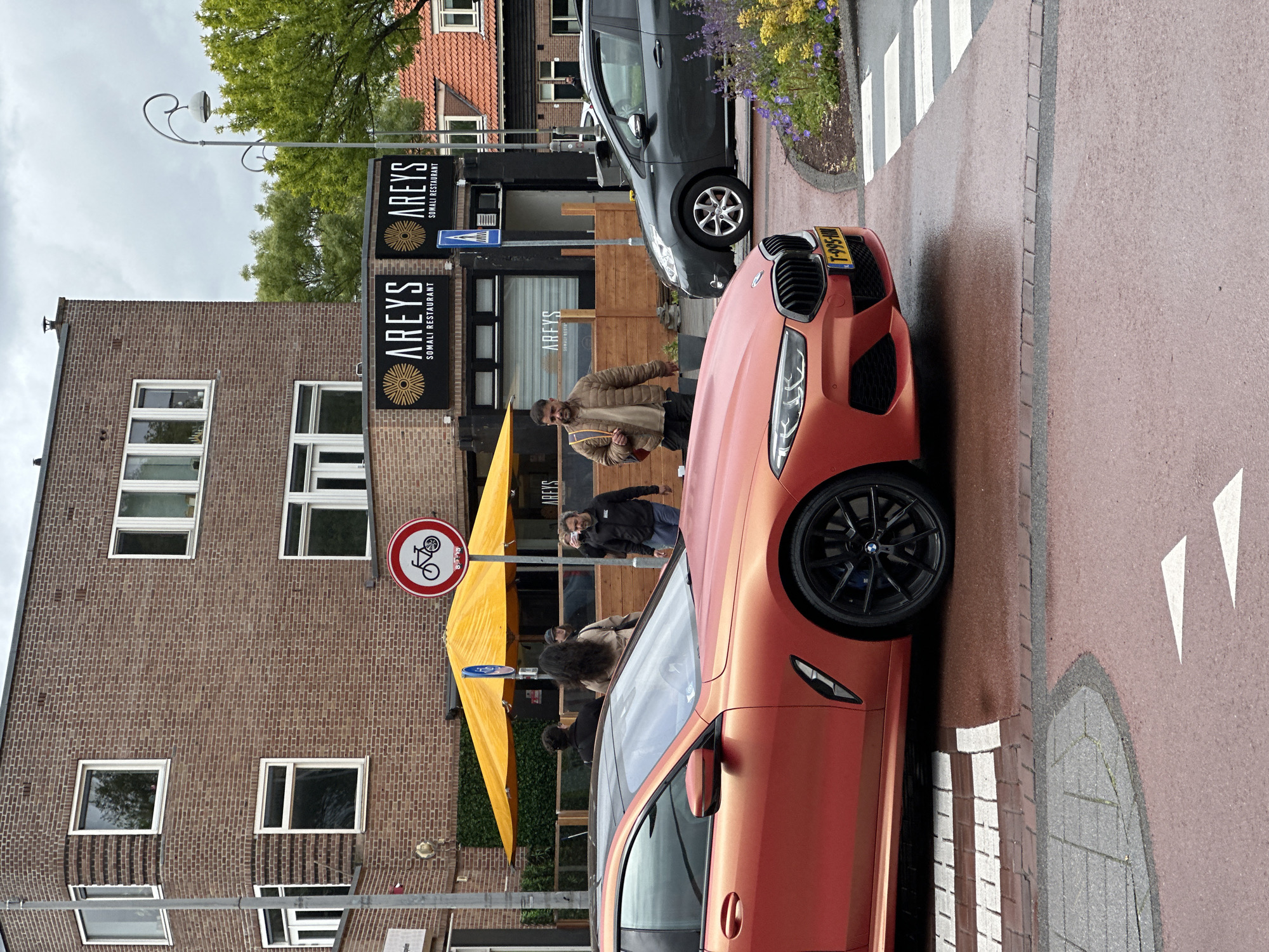 The image shows a street scene in Amsterdam, Netherlands. A coral-colored BMW is parked diagonally across a pedestrian crosswalk. Behind the car is a gray compact car. Pedestrians are visible near a building with signs indicating "Areys Somali Restaurant." A yellow awning is partially visible. A street sign indicates no bicycles allowed. The building is made of brick and has multiple windows. The sky is overcast.