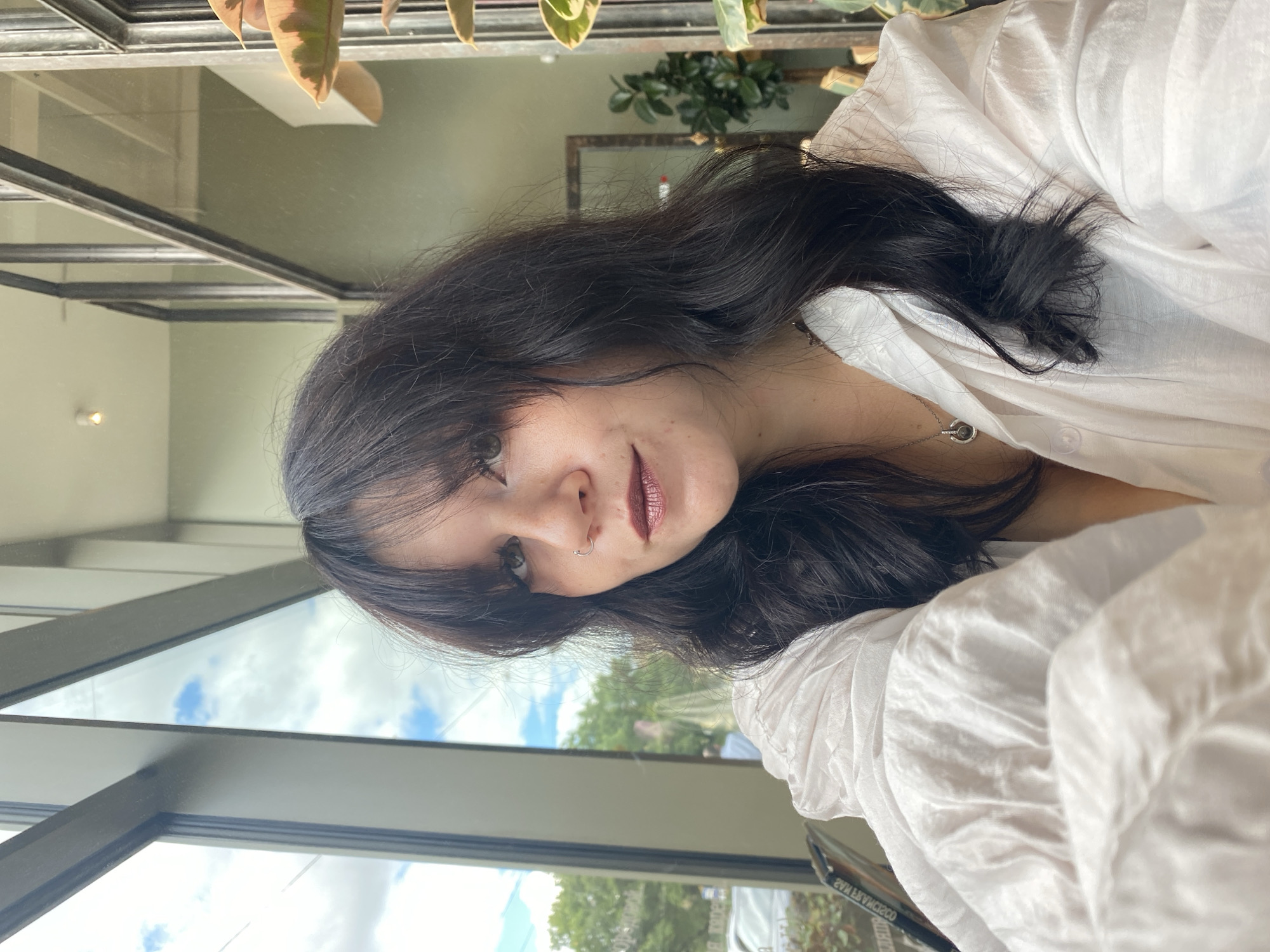 A young woman with dark wavy hair and bangs is pictured in a selfie taken indoors, likely in a cafe or restaurant, with large windows in the background. She is wearing a white collared shirt with a subtle sheen and a silver pendant necklace. Her right side is facing the camera, and she has a nose ring and dark lipstick. Through the window, glimpses of the sky, trees, and a street can be seen. The interior appears to have a modern design with metal framing and plants. The image does not provide any specific indicators of the location being Saint Petersburg, Russia.