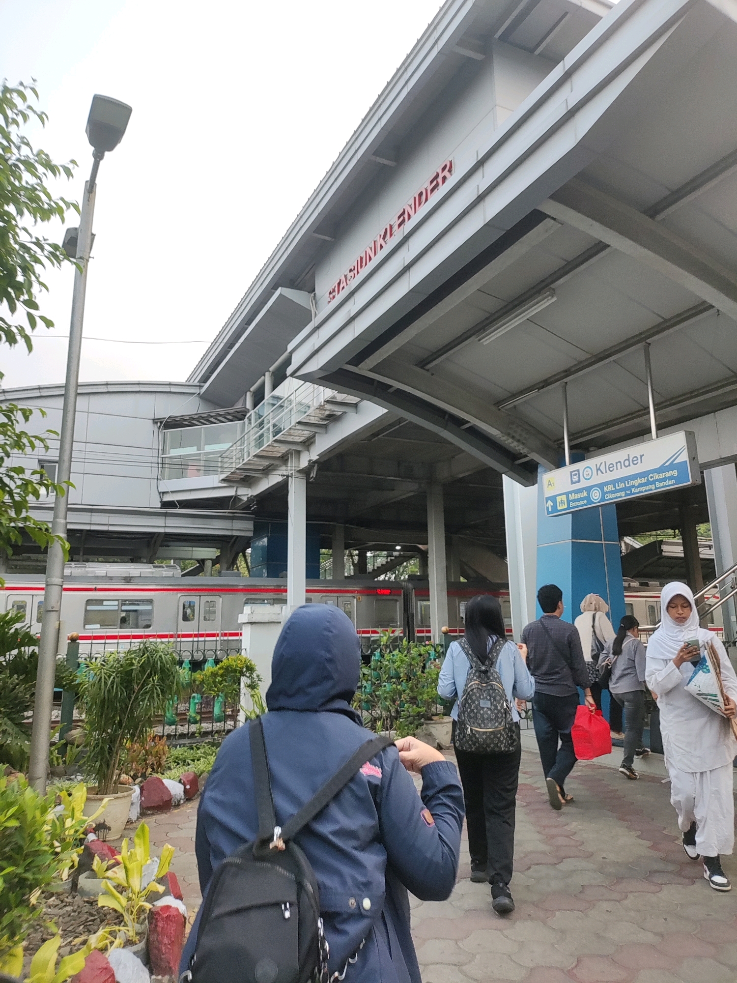 The image captures the exterior of the Klender Station in East Jakarta, Indonesia. The station's architecture features a modern, multi-level design with a prominent overhanging roof structure. Red lettering spelling "STASIUN KLENDER" is visible on the upper facade of the building. A train is parked on the tracks, identifiable by its gray and red color scheme. Several individuals are walking on the paved walkway leading towards the station. One person in the foreground wears a blue hooded jacket and a black backpack. Other pedestrians are dressed in casual attire, with some carrying bags. Green foliage and decorative plants are present on the left side of the walkway, bordering the station grounds. The sky is overcast.