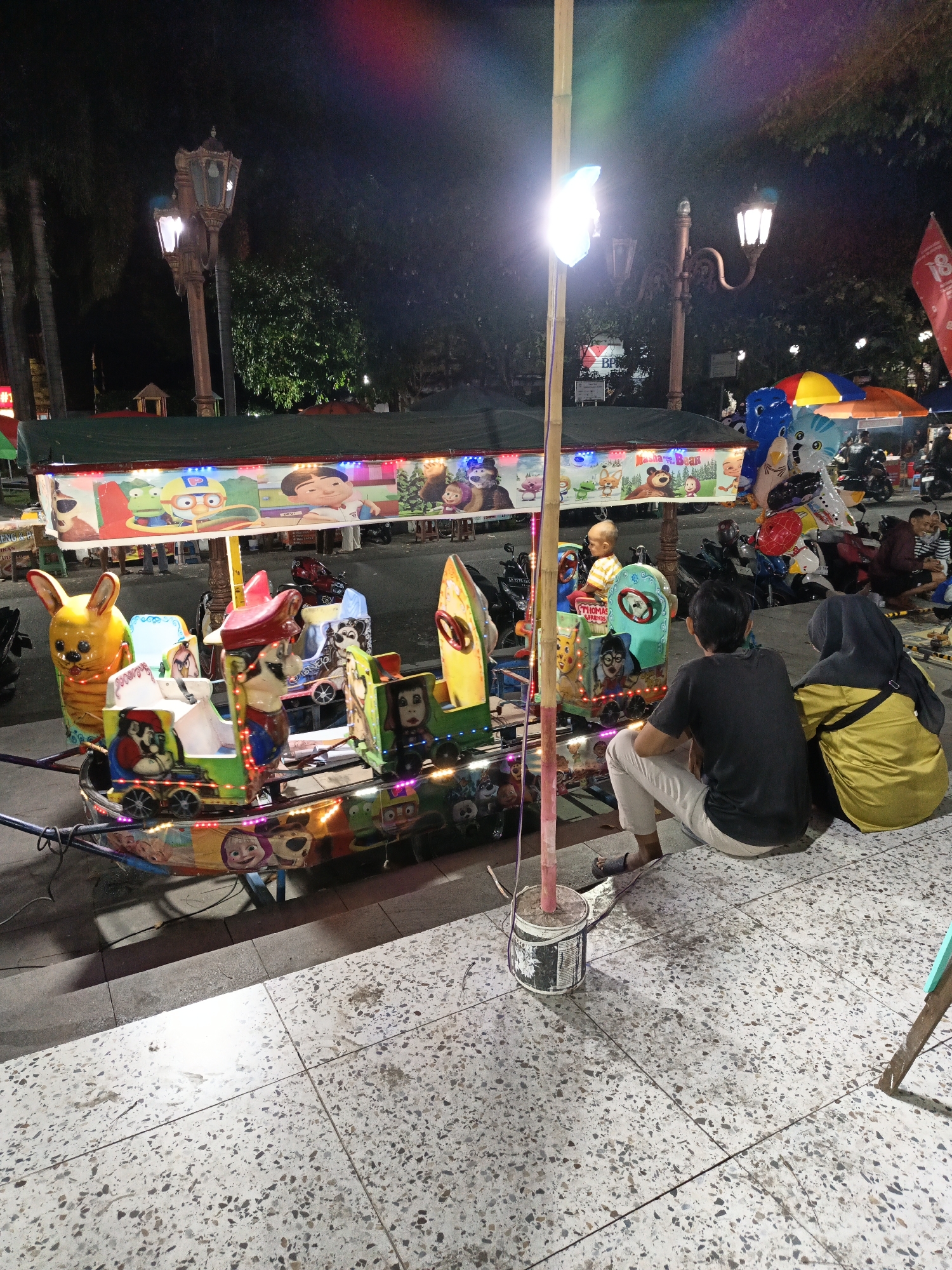 A brightly lit amusement ride for children is the central focus of this night scene, likely at a public fair or park in Klaten, Indonesia. The ride is decorated with cartoon characters and colorful lights. In the foreground, two individuals, a man and a woman, are seated on the ground, facing the ride. The man is wearing a black t-shirt and light-colored pants, while the woman is in a yellow hijab and jacket. Behind them, other people and motorcycles are visible, suggesting a lively atmosphere. The environment is paved with speckled tiles, and ornate street lamps stand in the background, illuminating the area with their warm glow. Trees and foliage are also present, adding to the outdoor setting. The overall impression is one of a vibrant evening entertainment activity.