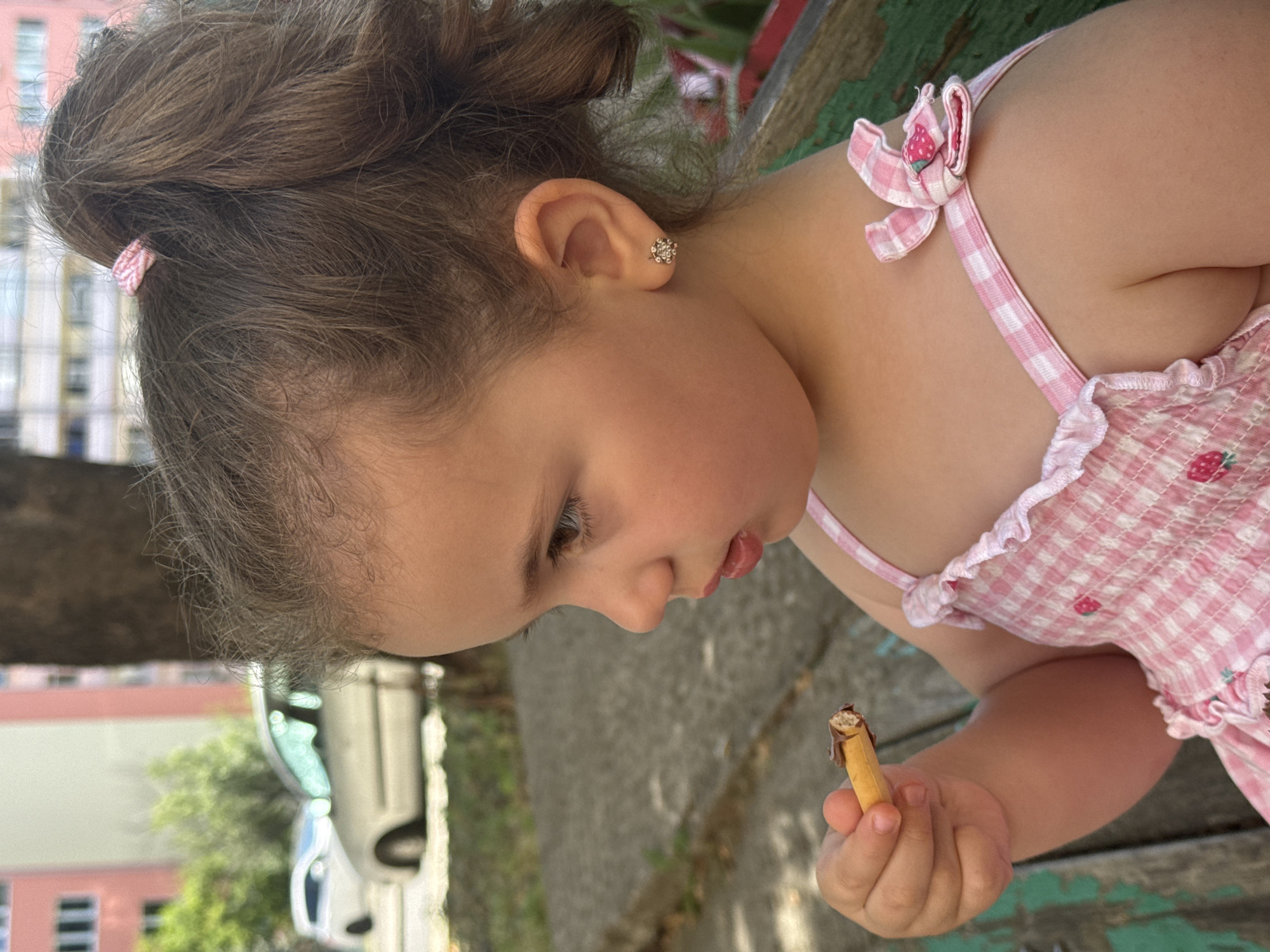 A young girl with brown hair in a ponytail, wearing a pink checkered dress with strawberry accents, is holding a chocolate-covered snack. She has a small, flower-shaped earring in her left ear. The girl is seated outdoors, with a blurred background that includes trees, buildings, and cars, suggesting an urban park or residential area. The scene appears to be captured on a sunny day.