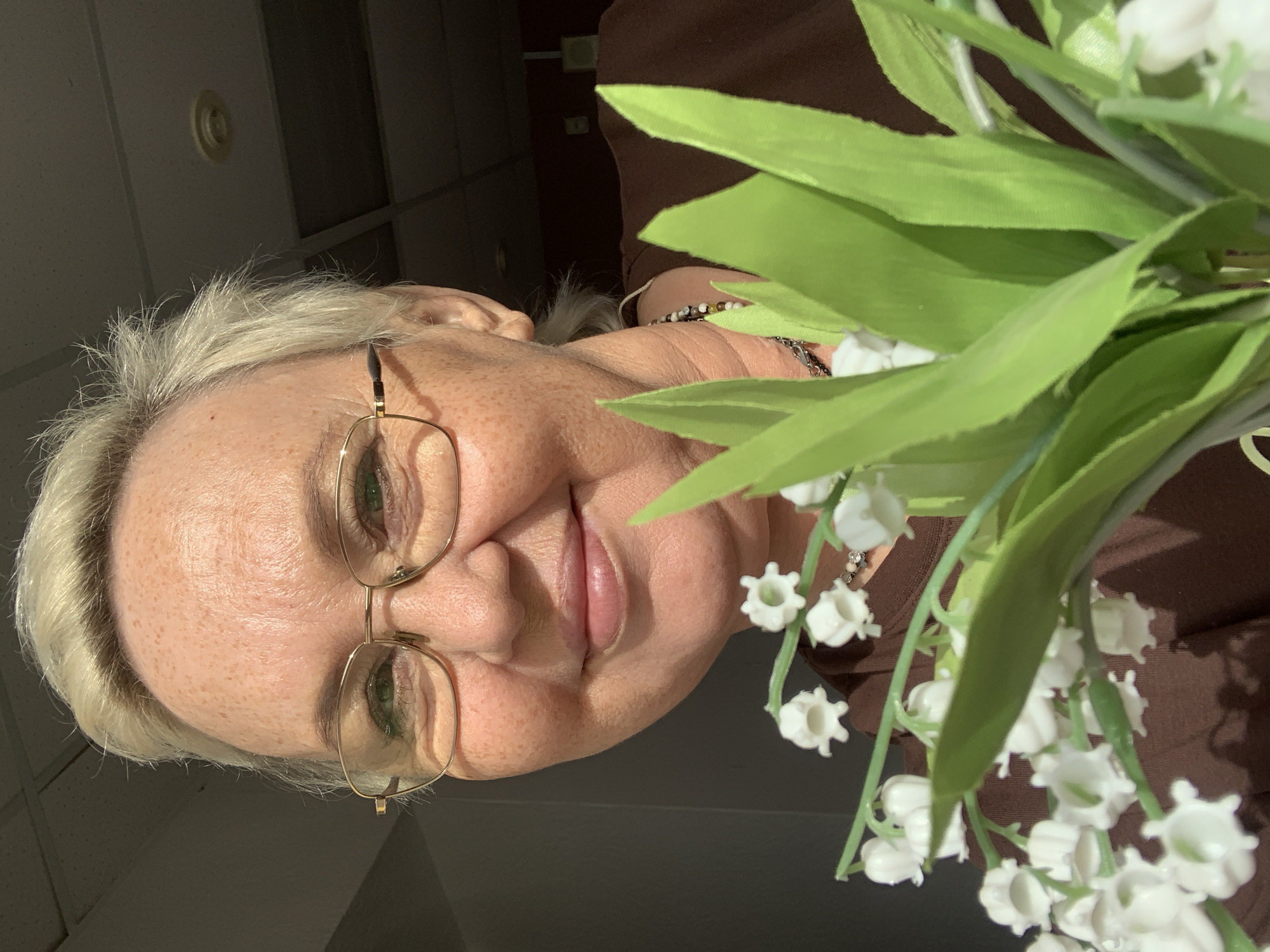 An outdoor, close-up, medium shot captures a Caucasian woman with blonde hair and fair skin, speckled with freckles. She is wearing gold-rimmed glasses and a brown shirt, and she is smiling as she holds a bouquet of white lilies of the valley with green leaves. The background is blurred, with a ceiling that appears to be from an indoor setting. The lighting suggests it might be daytime. The overall setting is ambiguous and does not provide clear indicators for a specific location within Chaikovsky, Russia, or any other city.