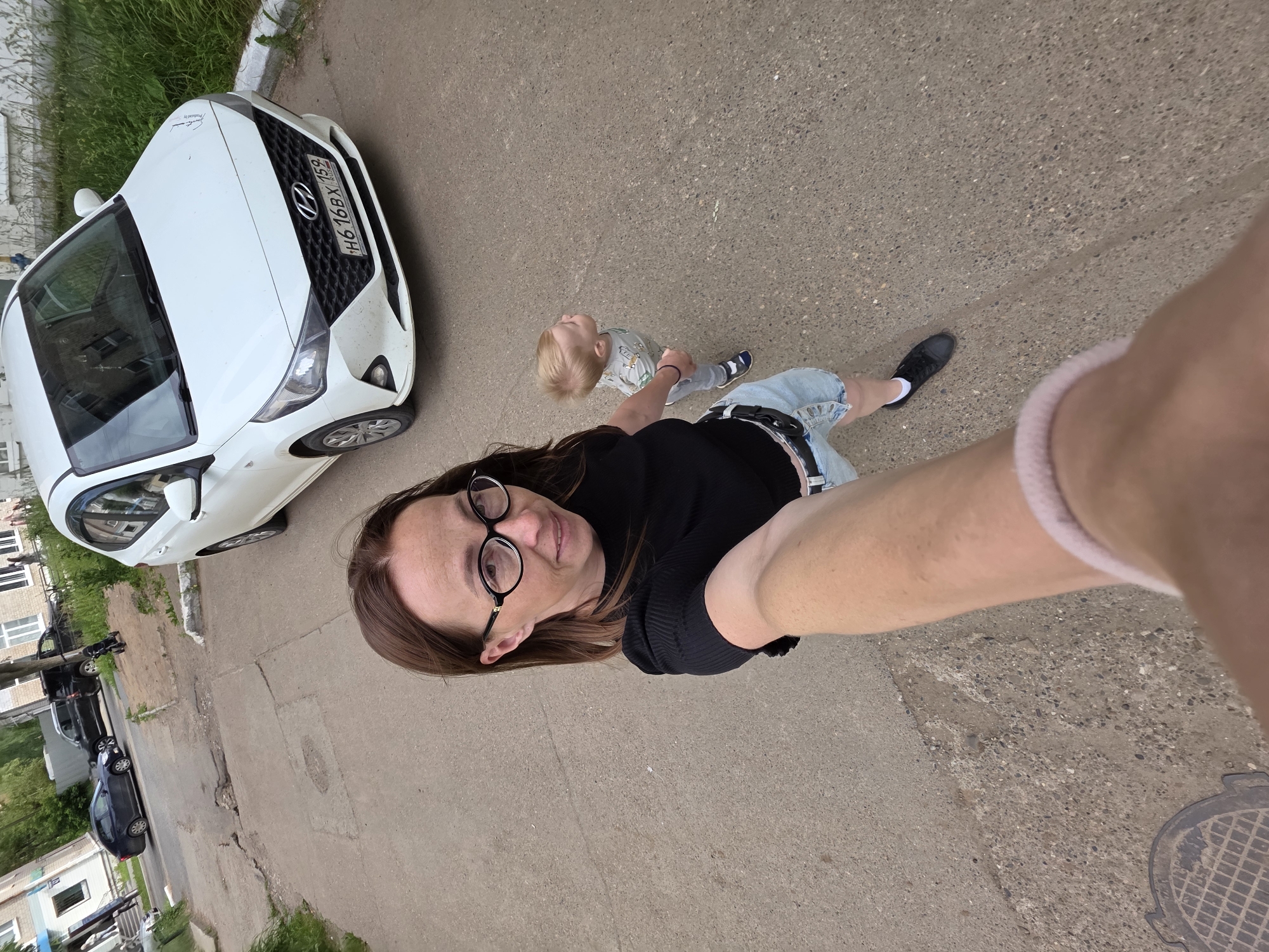 A person is taking a selfie outdoors with a young child standing nearby. The person in the foreground, wearing glasses and a black top, is holding the camera with their right arm extended. The child, with blonde hair, is wearing a grey t-shirt and denim shorts. They are standing on a paved surface, likely a sidewalk or road, with a white car parked to the left. The background shows more pavement and some greenery, suggesting an urban or suburban environment. Further down the paved area, several other parked cars are visible. The image appears to be taken during daylight, and the weather seems to be overcast. The license plate on the white car is visible and reads "H616BX 152". The scene is set in Chaikovsky, Russia. The overall mood seems casual and everyday.