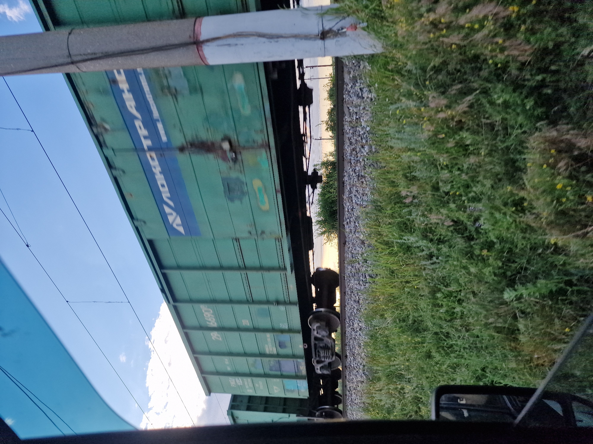 The image shows a side view of a green freight train moving along a railway track. The train cars have a textured, ribbed surface and some visible rust. A blue stripe runs along the side of the train cars with white lettering. The inscription "ЛОКО ТРАНС" is clearly visible on one of the cars. Below that, a number "29 1890" is visible.
The train is situated next to a grassy embankment with some yellow wildflowers. In the background, trees and a body of water can be seen, suggesting a rural or semi-rural setting. The sky is blue with some scattered white clouds. Overhead, power lines stretch across the frame. The perspective suggests the photo was taken from another moving vehicle, possibly a car, as a portion of a car's windshield and a side mirror are visible in the foreground.
The lighting indicates it is either early morning or late afternoon, with a warm glow on the horizon. The overall mood is tranquil and observational. The location is identified as Kursavka, Russia. There are no people clearly visible in the image.