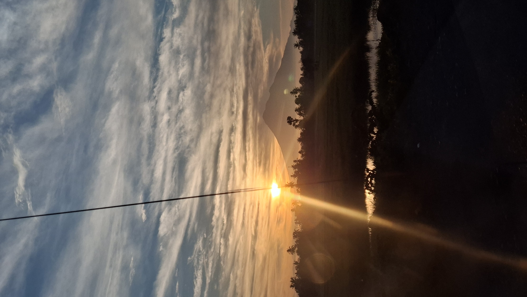 The image depicts a silhouetted landscape at sunrise, with a prominent mountain in the background. The sky is filled with wispy clouds that are illuminated by the rising sun, casting a warm orange and yellow glow. In the foreground, a thin, dark line, likely a power line, runs vertically through the frame. The middle ground features a silhouette of palm trees and other foliage, suggesting a tropical or subtropical environment. A body of water, possibly a rice paddy or a small pond, reflects the sunrise colors. The overall impression is one of a serene natural setting at the beginning of a new day. The location is identified as Aneukglee, Indonesia.