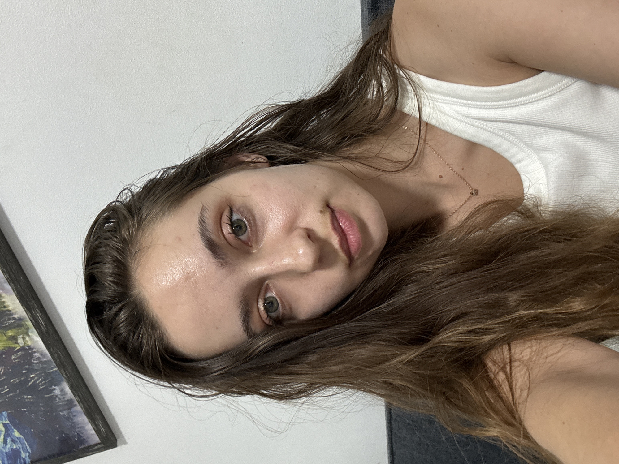 A close-up selfie of a young woman with light skin, brown hair, and green eyes. She is wearing a white tank top and a delicate gold necklace. The background includes a white wall and a framed picture. The location is unknown, but the subject specified Ramat Gan, Israel, which cannot be verified within the context of the image.