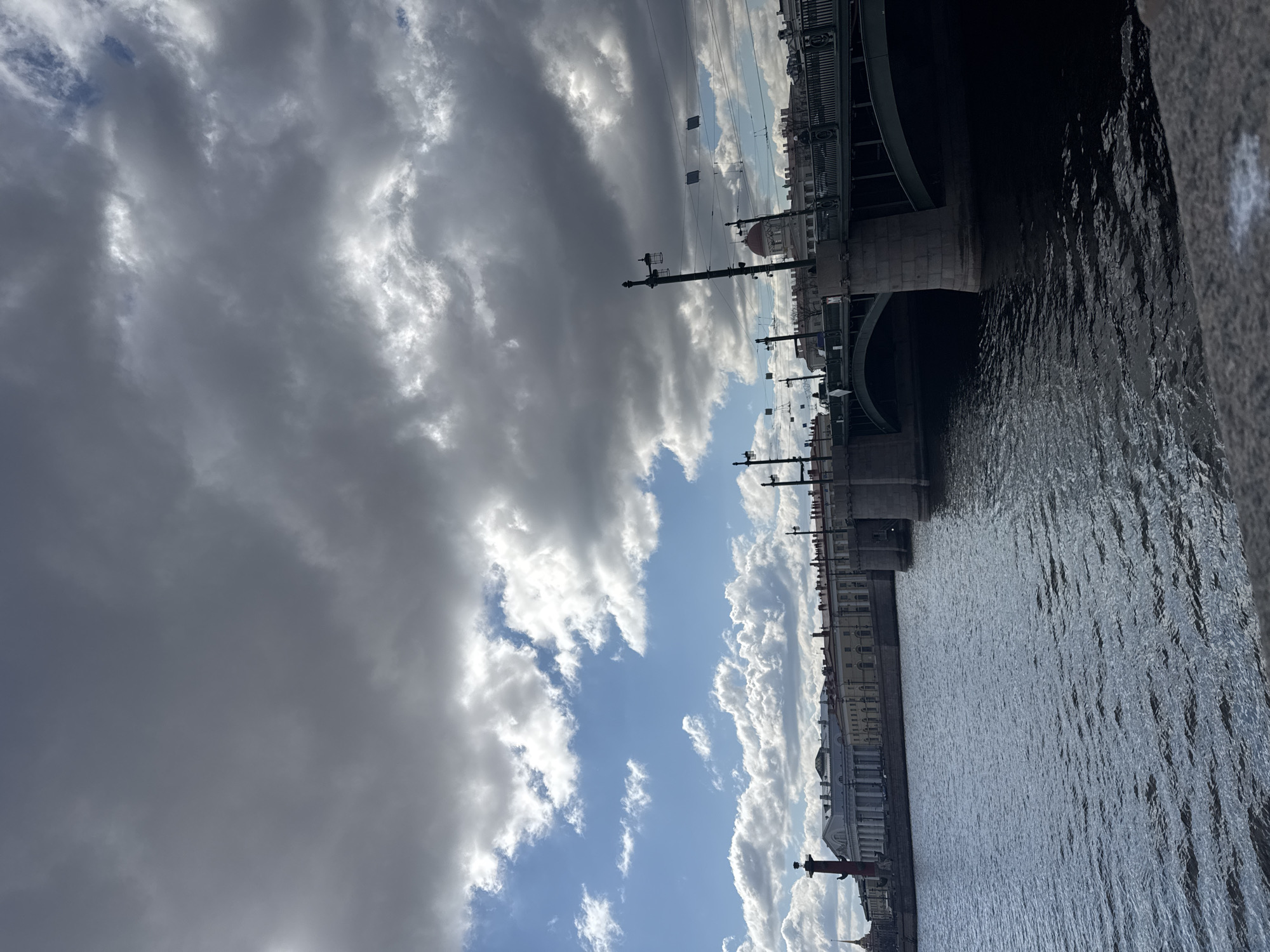 The image captures a panoramic view of the Neva River in Saint Petersburg, Russia, under a dramatic sky. The scene is dominated by a wide expanse of water on the right, showing ripples and reflections. A large, dark-colored bridge, likely a drawbridge, spans the river in the middle ground, featuring arched supports and decorative railings with lampposts.
Along the far bank of the river, a lengthy stretch of classical architectural buildings with columns and ornate facades lines the embankment. In the distance, towards the left, a prominent red Rostral Column with a sculpture on its top is clearly visible, a significant landmark of the city. Further down the river, another similar column can be faintly seen.
The sky above is a striking element, with dark, heavy storm clouds occupying a large portion of the frame, contrasting sharply with patches of bright blue sky and fluffy white cumulus clouds. The lighting suggests a dramatic day, possibly with intermittent sunlight breaking through the clouds. No people or specific activities are discernible, and no visible text is present. The overall impression is one of grand urban architecture set against a powerful, dynamic natural backdrop.