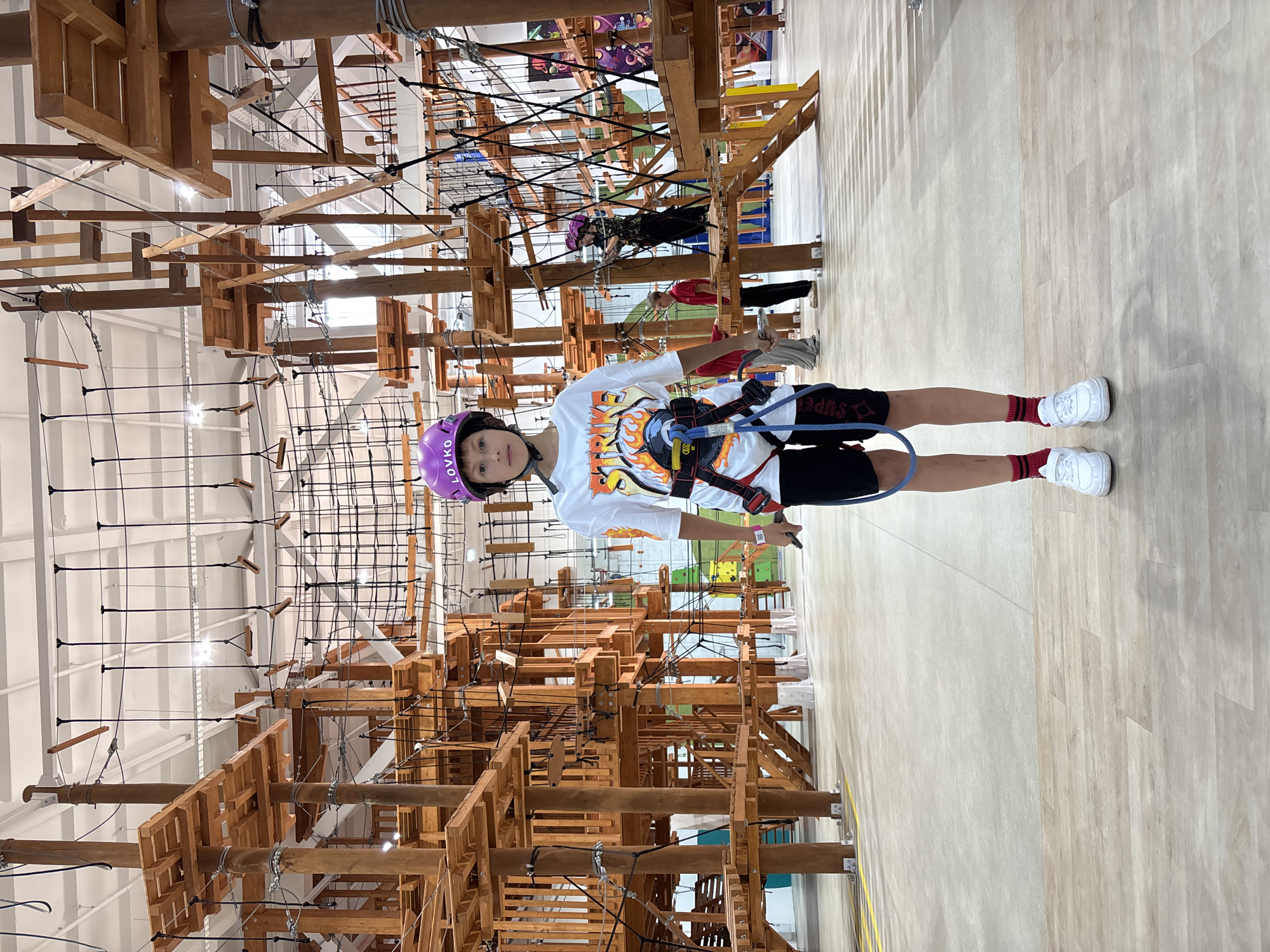 A young boy stands on a wooden floor in an indoor adventure park. He is wearing a purple helmet, a white t-shirt with flames and the word "STRIKE" on it, black shorts, red and black striped socks, and white sneakers. He has a harness on, with safety lines attached. Behind him, there is a complex structure of wooden platforms, ropes, and nets, typical of a ropes course. Several other people are visible in the background, also engaged in the activity. The park appears to be well-lit, with a high ceiling and industrial lighting. The overall impression is of an active, recreational environment. This location is the "Lovko" adventure park in Orenburg, Russia.