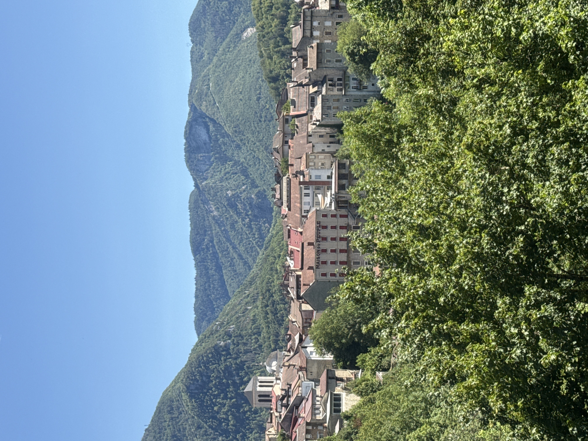 This image captures a view of the city of Saint-Claude, France, nestled amidst lush green mountains under a clear blue sky. The foreground is dominated by the dense foliage of large trees, obscuring the lower portion of the scene.
Rising above the trees, a cluster of buildings forms the heart of the city. The architecture suggests a historic European town, with many structures featuring terracotta tiled roofs. Prominent amongst these buildings is a church with a distinct tower and dome, its stone facade visible. Another notable building, a multi-story structure with a light-colored facade and red-shuttered windows, bears the inscription "MAISON DU PEUPLE". The buildings are packed closely together, indicative of an urban environment.
The middle ground and background are characterized by the rolling, densely forested mountains that encircle the city. The slopes are covered in a thick carpet of trees, with visible textures and layers indicating varying terrain and tree density. The clear, cloudless sky provides a bright, sunny backdrop to the entire scene. There are no people visible in the image.