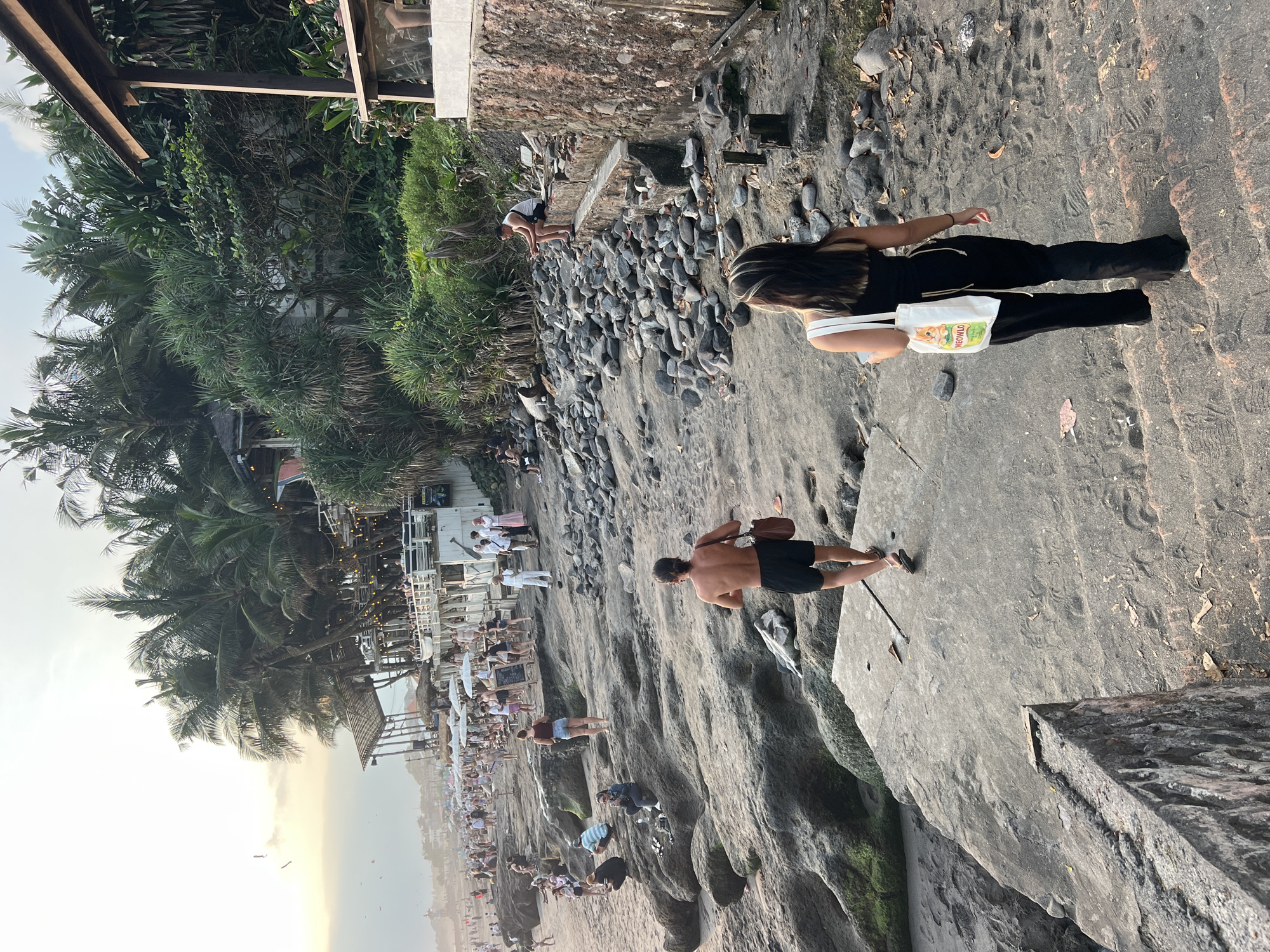 This is an eye-level, medium shot of a beach scene in North Kuta, Indonesia, during a late afternoon. The foreground shows a concrete walkway with a woman in black clothing and a white tote bag with a cat image walking away from the camera up a set of stairs. To her left, a shirtless man in black shorts with a brown bag slung over his shoulder is walking on a rocky surface, also moving away from the camera. In the background, a crowded beach with palm trees and several open-air restaurants with white umbrellas and string lights is visible. The ocean is not visible.