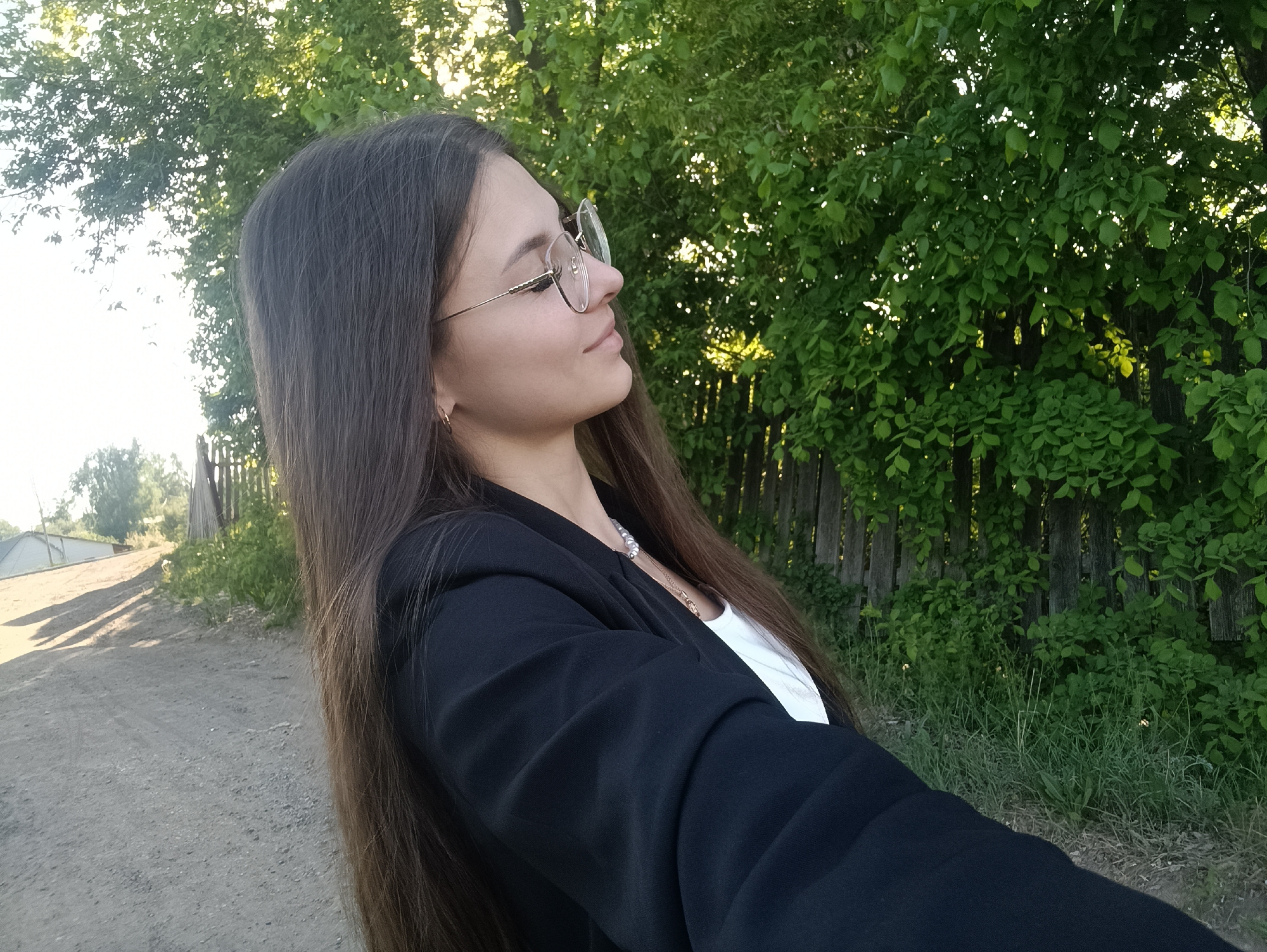 The image shows a woman in a black blazer and glasses, standing outside. The background includes trees and a wooden fence. She appears to be on a dirt road. The exact location within the city of Unknown, Unknown is not discernible from the image.