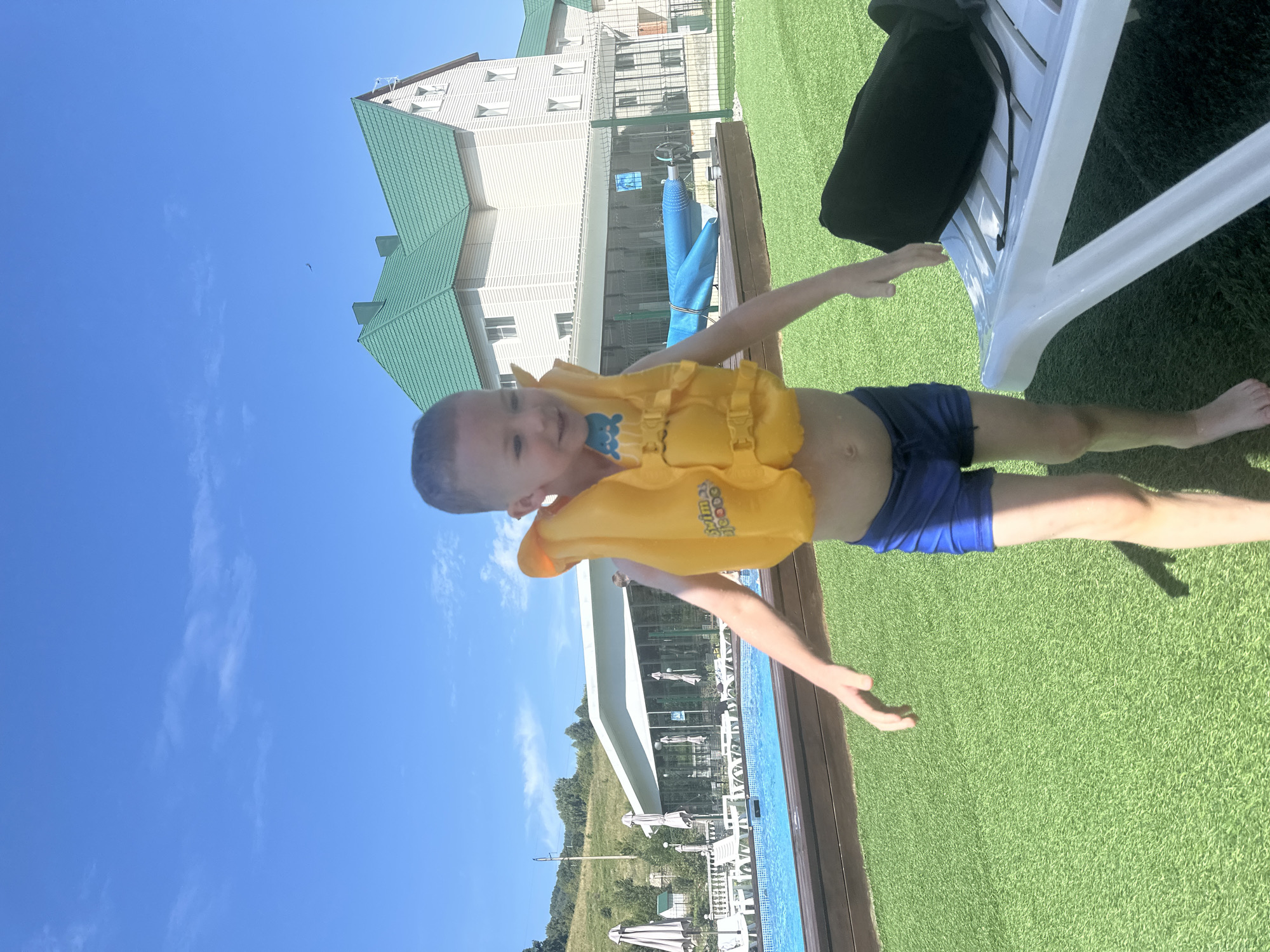 A young boy, wearing a bright yellow life jacket and blue swim trunks, stands on artificial turf near a swimming pool. The boy is smiling and appears to be in motion. In the background, there is a large building with a green roof, lounge chairs, and umbrellas. The setting appears to be a recreational facility in the city of Danilovka (Altai), Russia, on a sunny day with a clear blue sky.