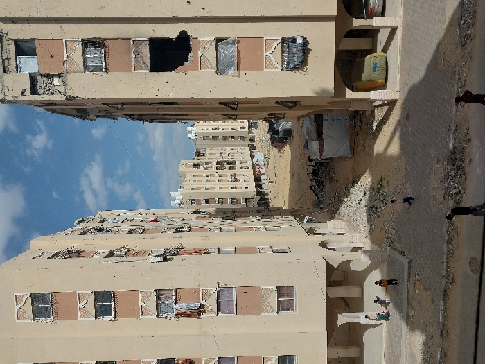 This image depicts a scene within Khan Yunis, Palestinian Territory, characterized by heavily damaged multi-story residential buildings under a partly cloudy sky. Two prominent light-tan apartment blocks, each displaying decorative patterns, frame a central unpaved area. Both foreground buildings exhibit extensive damage, including numerous impact marks, shattered windows, missing panes, and exposed interior sections; some window openings are covered with tarps or fabric. On the ground level of the building on the right, a vehicle obscured by a yellow tarp is parked within an arched recess, alongside a partially visible silver car. Under the arched ground floor of the left building, three children are standing. The central ground area is an expanse of sand and rubble. A child in a blue shirt walks across this unpaved space. Further back, several makeshift shelters constructed from tarps are visible, interspersed among additional multi-story apartment complexes that also show signs of wear and damage. In the immediate foreground, two individuals are partially visible walking on a paved section. The scene is illuminated by daylight, casting distinct shadows.