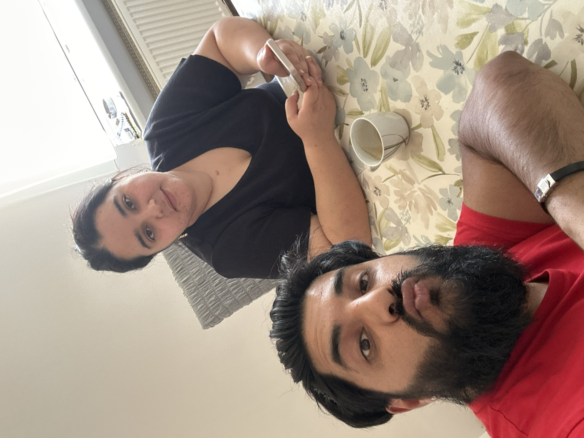 The image shows two people, a woman and a man, lying down. The woman is wearing a black t-shirt and is holding a smartphone, appearing to be engaged with it. She has dark hair and is looking towards the camera. To her right, a white mug with some liquid, possibly coffee or tea, is visible on a patterned surface that resembles a tablecloth or bedspread. The man, wearing a red t-shirt, is lying next to her. He has a beard and is also looking towards the camera. His arm, adorned with a black bracelet with a silver buckle, is visible. The background suggests an indoor setting, possibly a bedroom or living room, with a radiator visible in the upper left corner. The lighting indicates it is daytime. The overall impression is a casual, relaxed moment shared between the two individuals, possibly at home.