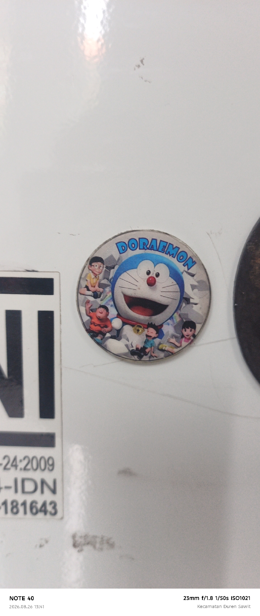 A round, decorative sticker featuring the cartoon character Doraemon and several other animated figures is affixed to a white, glossy surface. The sticker shows Doraemon in the center, with Nobita, Shizuka, Gian, and Suneo surrounding him. To the left of the sticker, a portion of a black and white label with text and symbols is visible. The label contains the letters "NI" in large, bold font, followed by smaller text including "-24:2009", "-IDN", and "-181643". The white surface exhibits some minor smudges and scratches. The bottom of the image displays text overlays indicating "NOTE 40", "2026.08.26 13:41", "23mm f/1.8 1/50s ISO1021", and "Kecamatan Duren Sawit", the latter being the administrative district of East Jakarta, Indonesia.