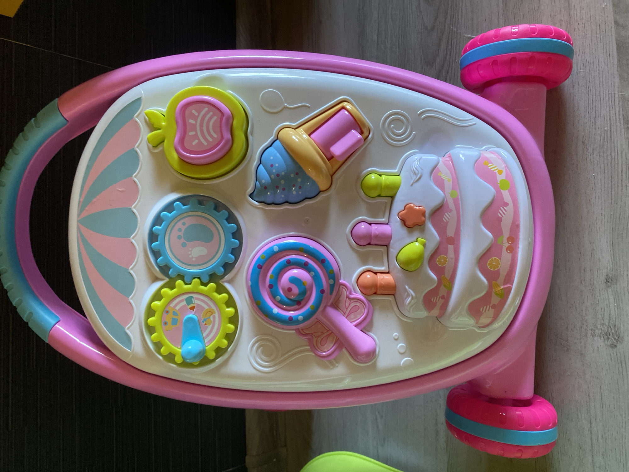 A pink and teal baby walker with various toys is situated on a wooden floor. The walker features a large handle at the top, with colorful interactive elements like gears, an apple-shaped button, a lollipop, and an ice cream cone. A tiered cake design is prominent in the lower section, adorned with colorful decorations and what appear to be toy candles. Two pink wheels with teal and pink stripes are attached to the base. The background consists of a dark, possibly wooden, surface and a lighter wooden floor. The image is taken from a slightly elevated, front-facing angle. The location is not determinable from the provided image.