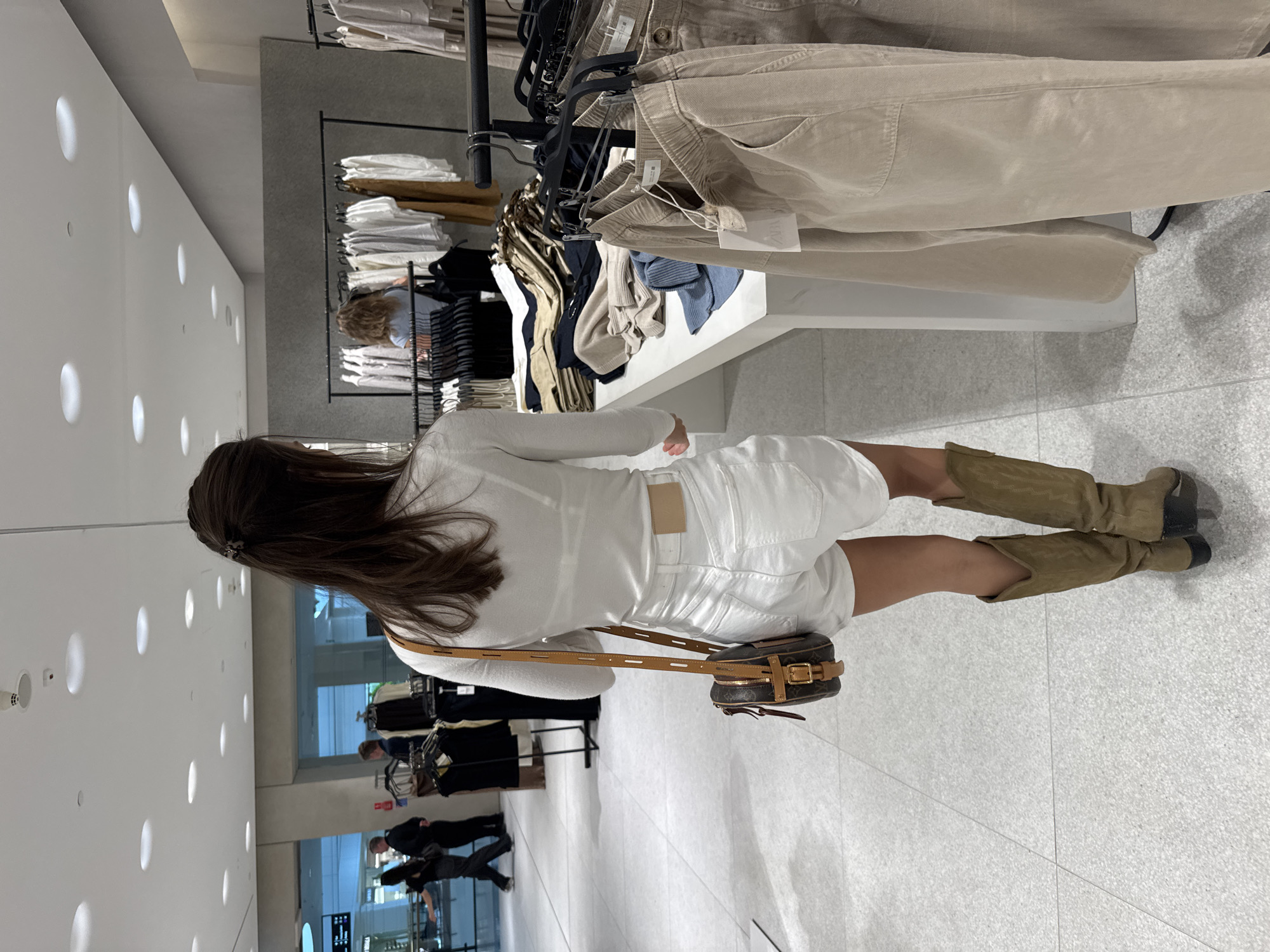 A woman wearing a white top and shorts with knee-high brown boots is shopping in a clothing store. She is looking at a rack of neutral-colored pants. Other racks of clothing and shoppers can be seen in the background. The store appears to be modern with white walls and bright lighting. The location is in an unidentified city.