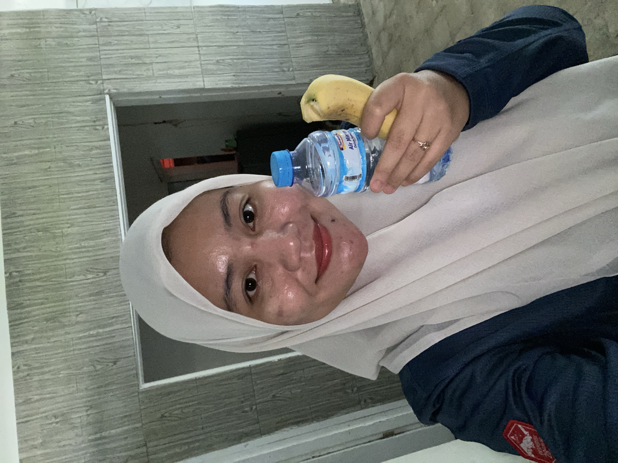 In Banjarbaru, Indonesia, a person wearing a white hijab and a navy blue jacket, adorned with a red and white patch on the left sleeve, is photographed from the chest up. They are smiling and holding a small bottle of "Air Minum" (drinking water) in their right hand, with a banana in the same hand, positioned to the right of the water bottle. The background features a textured wall with a wood-like pattern and an open doorway leading into a dimly lit interior. The person has visible skin imperfections on their face and wears subtle makeup. A ring with two small diamonds is visible on their left ring finger.
