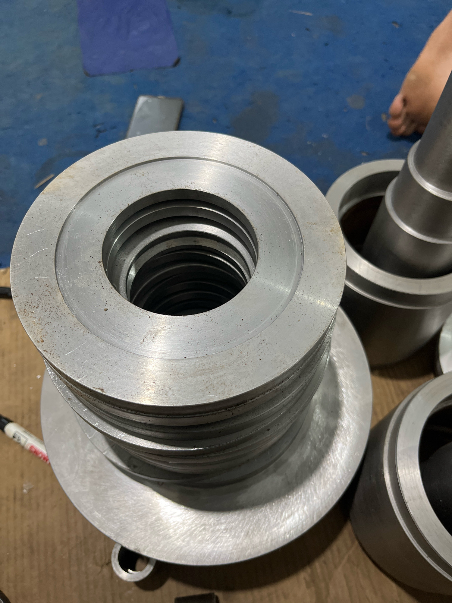 In a workshop setting, likely in Cimahi, Indonesia, a stack of identical metal rings with concentric grooves sits prominently in the foreground. The rings are made of a brushed metallic material, possibly aluminum or stainless steel, and show signs of industrial use with minor scuffs and dust. To the right of the stack, two cylindrical metal containers are partially visible, one appearing taller than the other and containing a darker object inside. In the background, a blue floor is visible, along with a dark rectangular object and a sliver of a person's foot. A white marker pen with red lettering is also present near the bottom left corner of the image, lying on a cardboard surface. The overall scene suggests a manufacturing or repair environment where these metal components are being stored or processed.