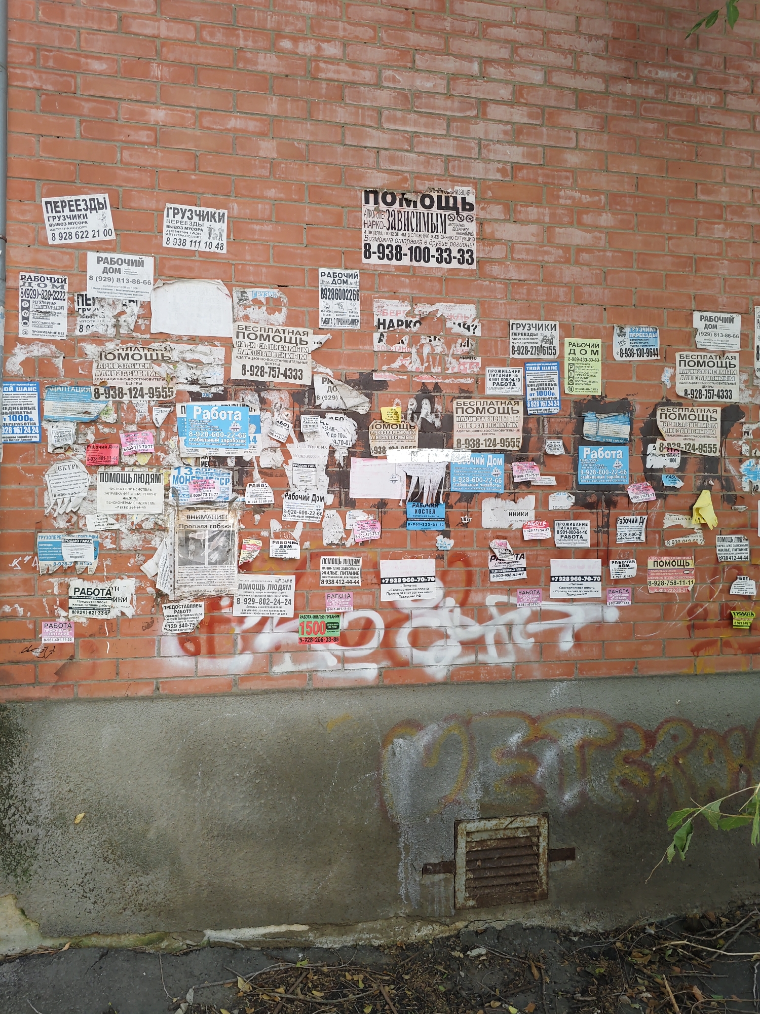 The image shows a brick wall in Rostov-on-Don, Russia, covered in numerous advertisements and flyers. These postings are primarily for services such as moving, moving and carrying services, job postings ("Rabochiy Dom," "Rabota"), and free assistance for drug addicts ("Besplatnaya Pomoshch"). Many of the flyers display phone numbers for these services. Some of the flyers are tattered and peeling, indicating age and exposure to the elements. There is also graffiti on the lower portion of the wall, obscuring some of the posters and the brickwork. The ground below the wall appears to be a mixture of concrete and dirt, with some debris and foliage.
