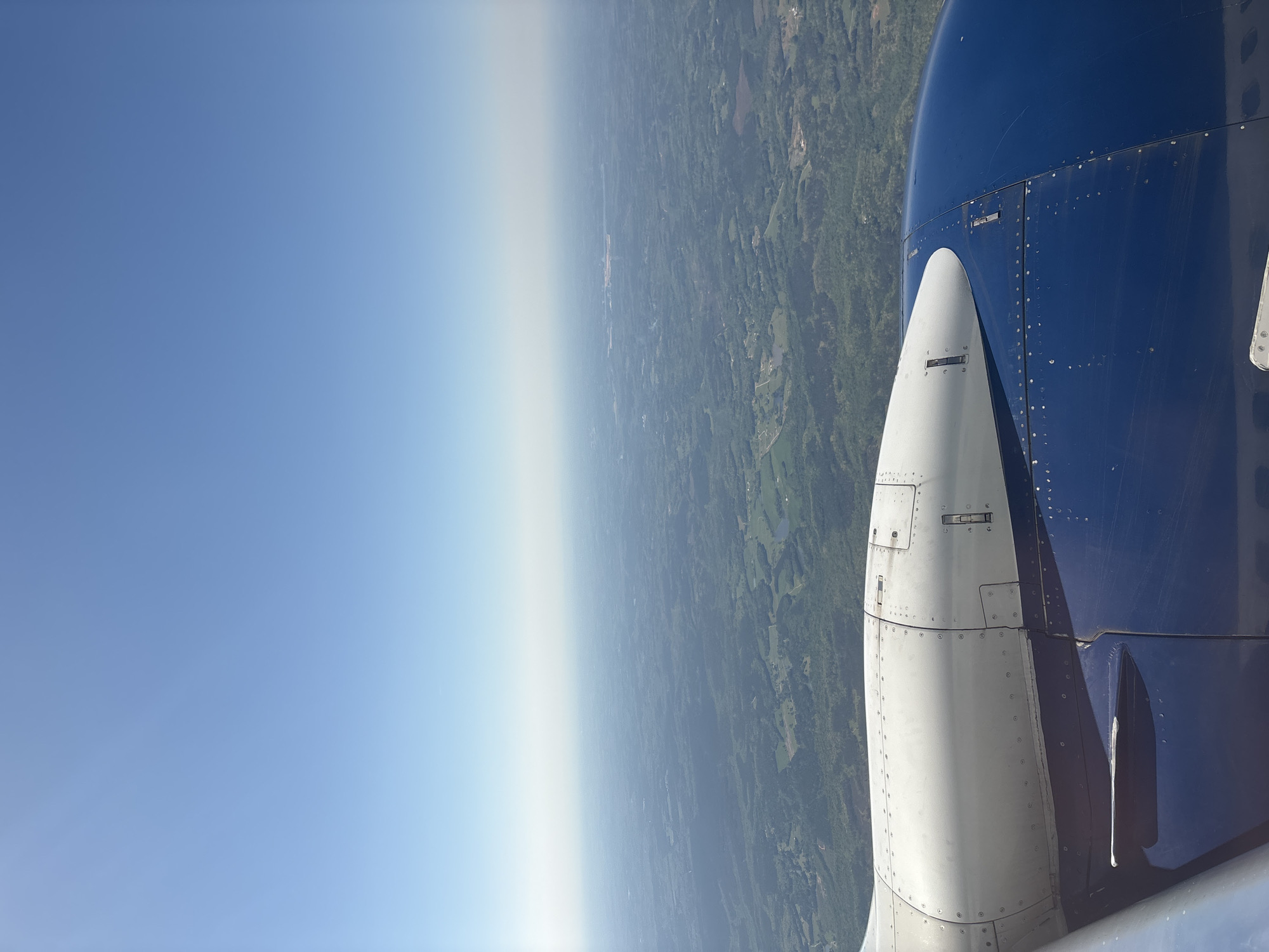 An aerial view from an airplane window. The window frame is on the right and consists of a blue and white part of the engine. Outside the window, a blue sky can be seen on the left and land on the right. The land consists of a mixture of forested areas and open fields. There are a few small bodies of water scattered across the landscape. The sky is clear blue with a slight haze near the horizon.