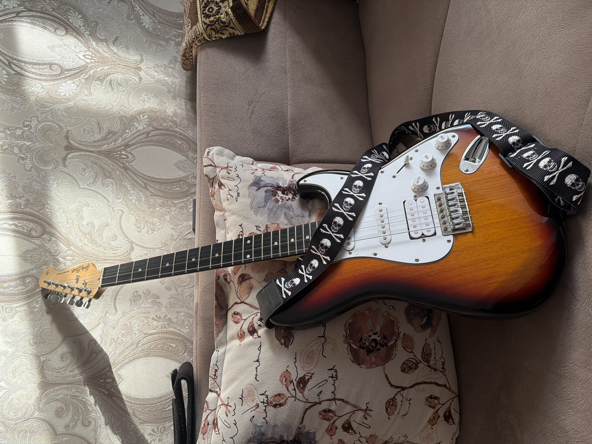 This media file features an electric guitar, likely a Fender Stratocaster due to its body shape and pickup configuration, lying on a couch. The guitar has a sunburst finish with shades of orange, red, and black. It is adorned with a black guitar strap decorated with a skull and crossbones pattern. The guitar is resting on a decorative pillow with a floral and script pattern. Behind the guitar and pillow, a textured brown couch is visible. The background shows patterned wallpaper with a textured, embossed design in light, neutral tones, and some sunlight is casting shadows on it. A hint of a patterned throw blanket is visible in the upper right corner. The scene appears to be indoors, possibly a living room, during the daytime given the natural light and shadows. There is no discernible activity taking place, suggesting the guitar is at rest. The overall mood is casual and relaxed.