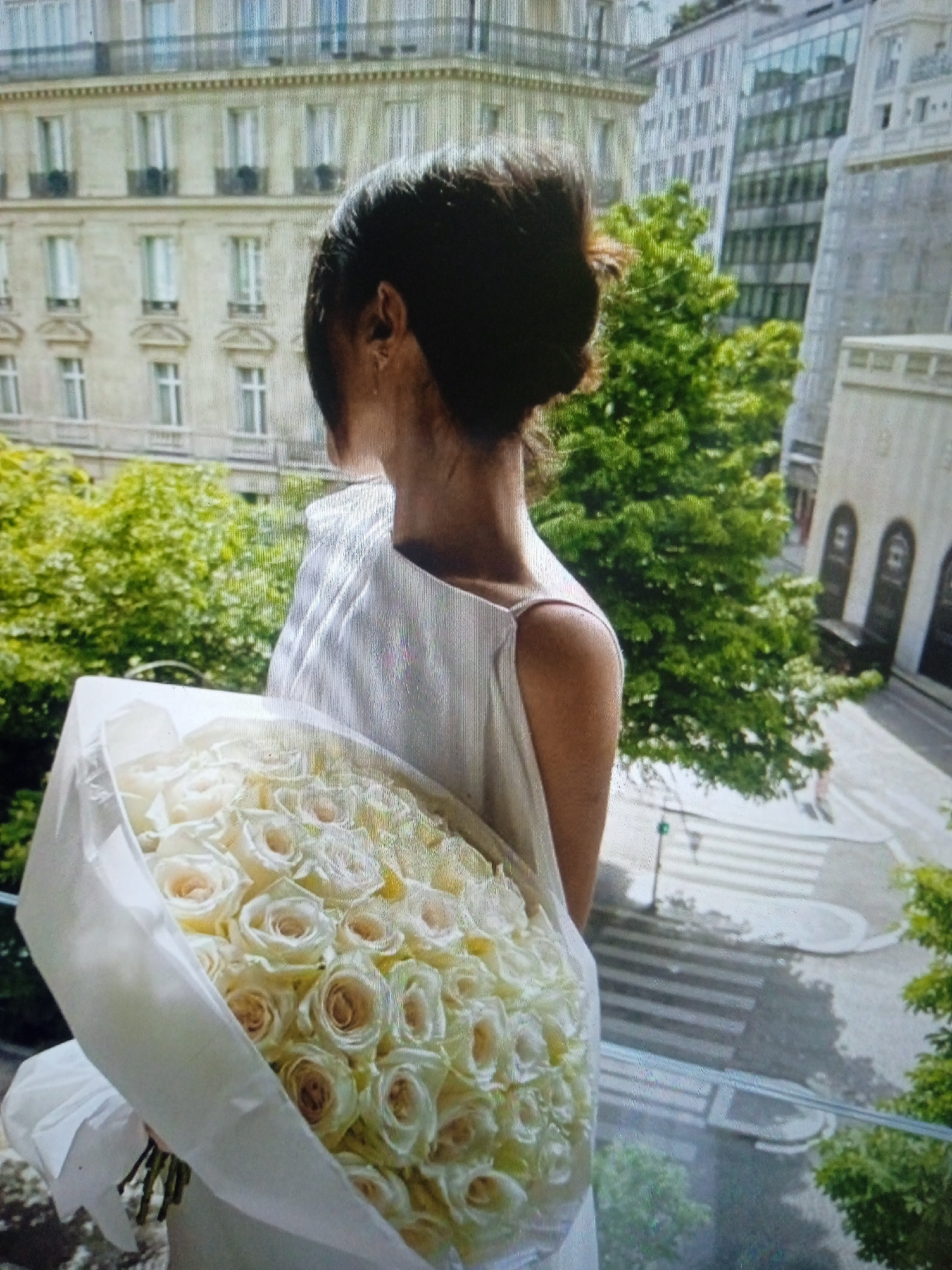 A person with dark hair styled in a bun is shown from the side, holding a large bouquet of light-colored roses. They are wearing a white dress with thin straps. The person appears to be standing on a balcony or terrace, overlooking a city street with buildings and trees. The architecture suggests a European city, possibly Paris given the style of the buildings. There is no indication of the location being Chernivtsi, Ukraine. The lighting suggests it is daytime, with natural light illuminating the scene.