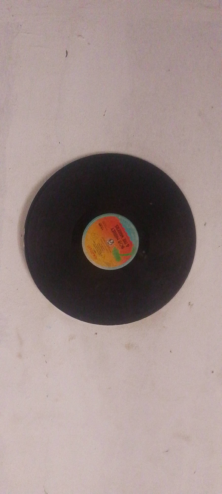 A black vinyl record with a colorful label is centered on a lightly speckled, light-colored surface. The label, with its orange, yellow, and blue sections, features text that appears to read "BOB MARLEY & THE WAILERS". The surface is textured and shows some minor imperfections and what might be dust specks. The lighting suggests an indoor setting, possibly a room in Jalingo, Nigeria, given the provided context. No people or actions are depicted.