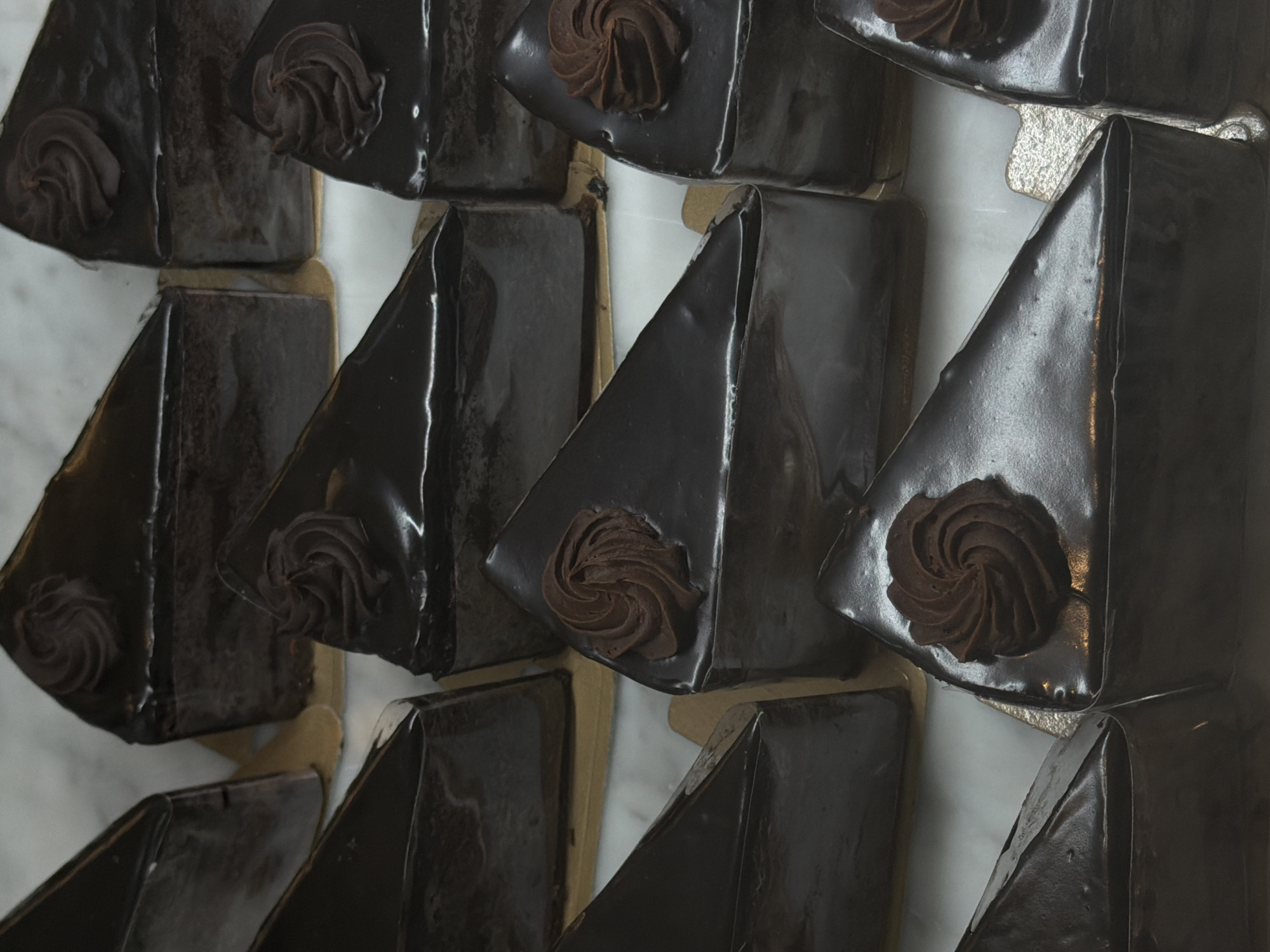 A high-angle, close-up shot reveals a selection of triangular chocolate cakes displayed in a likely bakery or pastry shop, potentially located in North Kuta, Indonesia. The cakes are arranged in a geometric pattern, with their glossy, dark chocolate glaze reflecting light. Each slice is adorned with a swirl of chocolate frosting in the center. The cakes rest on what appears to be a white, possibly marble or tiled surface. The lighting is somewhat dim, casting shadows and highlighting the shiny surfaces of the cakes. The scene suggests a focus on presentation and the arrangement of desserts for sale.