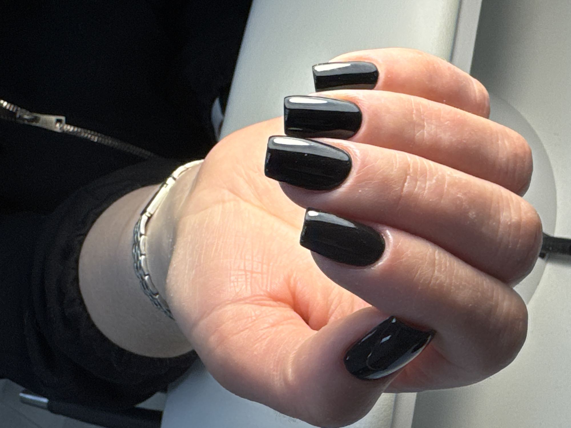 A light-skinned human left hand, located in the city of Chaikovsky, Russia, is depicted in a close-up, high-angle shot. The hand is positioned palm-up with fingers slightly curled, displaying manicured fingernails. All five visible nails are coated with a glossy black polish and exhibit a squoval shape. A silver-toned, linked bracelet consisting of rectangular segments is present on the wrist. The subject is wearing a black garment, likely a jacket, with a metallic zipper slider and teeth visible on the upper left portion of the image. The background is softly blurred, revealing a light-colored surface positioned to the right and below the hand, and a darker, indistinct area behind the arm. Natural light illuminates the hand and creates distinct reflections on the polished nails.