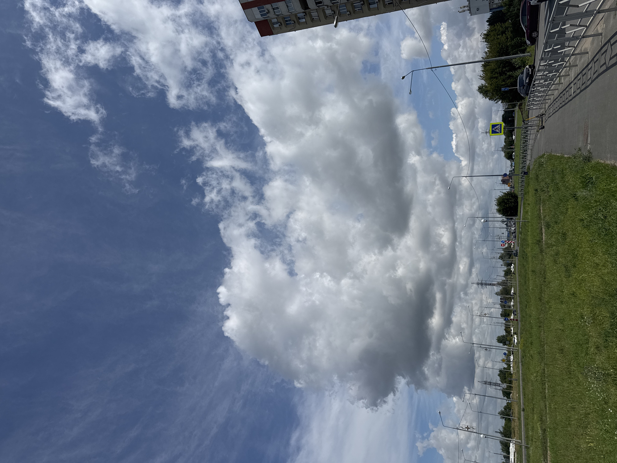This is a photograph taken from a sidewalk in Tolyatti, Russia, looking towards a road and a green field under a cloudy blue sky. The image captures a section of a residential building on the right, with multiple windows and balconies. A sidewalk with a metal railing lines the edge of the road, which has a pedestrian crossing sign and a parked electric scooter. Beyond the road, there is a stretch of green grass and trees, with a line of streetlights and power lines running parallel to the road. The sky is dominated by large, fluffy white clouds against a bright blue background, with some wispy cirrus clouds visible higher up. The overall impression is of a somewhat overcast but pleasant day in a suburban or semi-urban area.