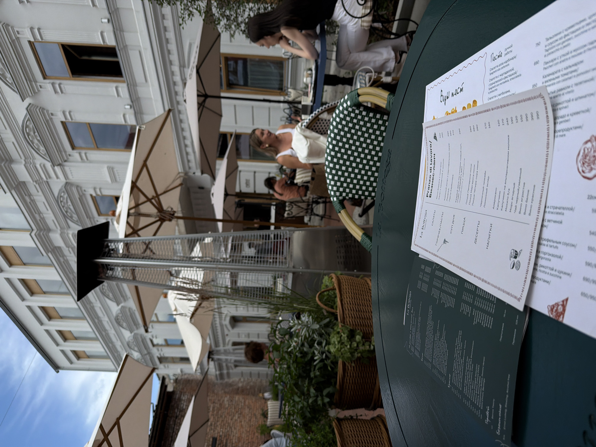 This is a photograph taken at an outdoor cafe, likely in Ufa, Russia, during the day. The scene is dominated by a large, dark green table in the foreground, upon which several menus are laid out. The menus are primarily in Russian and Italian, with some food and drink items listed, along with prices. Visible text includes "La Bottega," "Pranzo di lavoro/Бизнес-ланч," and various food categories like "САЛАТЫ" (Salads), "Паста" (Pasta), and "НАПИТКИ" (Drinks).
In the background, there is a white building with ornate architectural details and multiple windows. Several large outdoor umbrellas are visible, suggesting a sunny but potentially warm day where shade is needed. Two women are seated at a table under a checkered umbrella. One woman, wearing a white top, is looking to her left with a slight smile, appearing relaxed and engaged in conversation. Another woman, in a dark top, is also seated at the table, looking down. They appear to be enjoying a meal or drinks.
The overall atmosphere is casual and leisurely, typical of a cafe setting. Wicker baskets filled with greenery are placed around the seating area, adding a touch of natural decoration. The image is angled, with the building and sky appearing on the left side of the frame, creating a dynamic composition. The lighting suggests it is daytime, and the presence of umbrellas indicates fair weather.