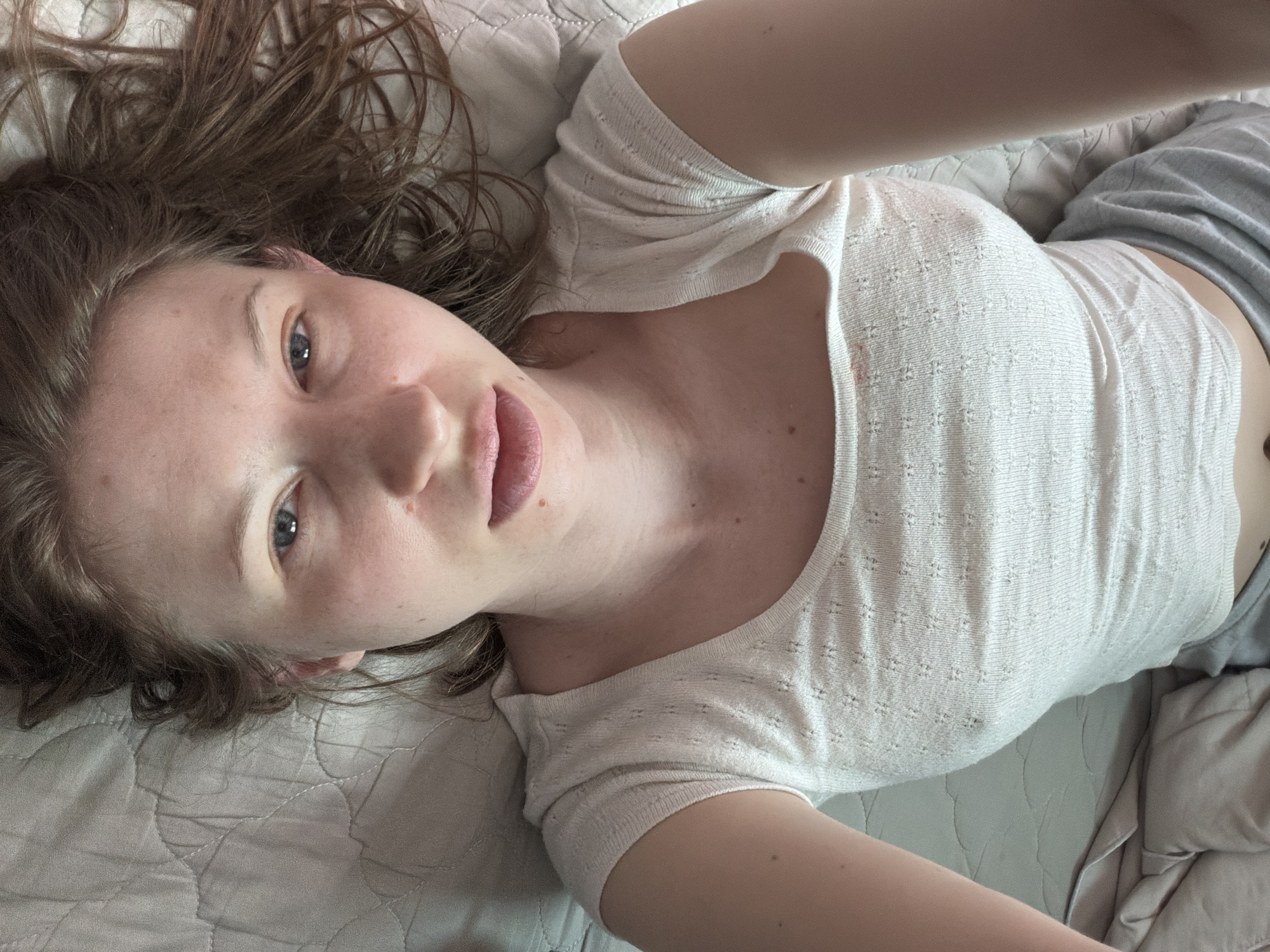 The image shows a person lying on a bed, upside down, facing the camera. The person is wearing a white, textured top and grey pants. Their arms are raised above their head, and their hair is spread out around them. The background is a light-colored quilted bedspread. The image does not contain any elements that would allow for the identification of a specific location in Dongducheon-si, South Korea.