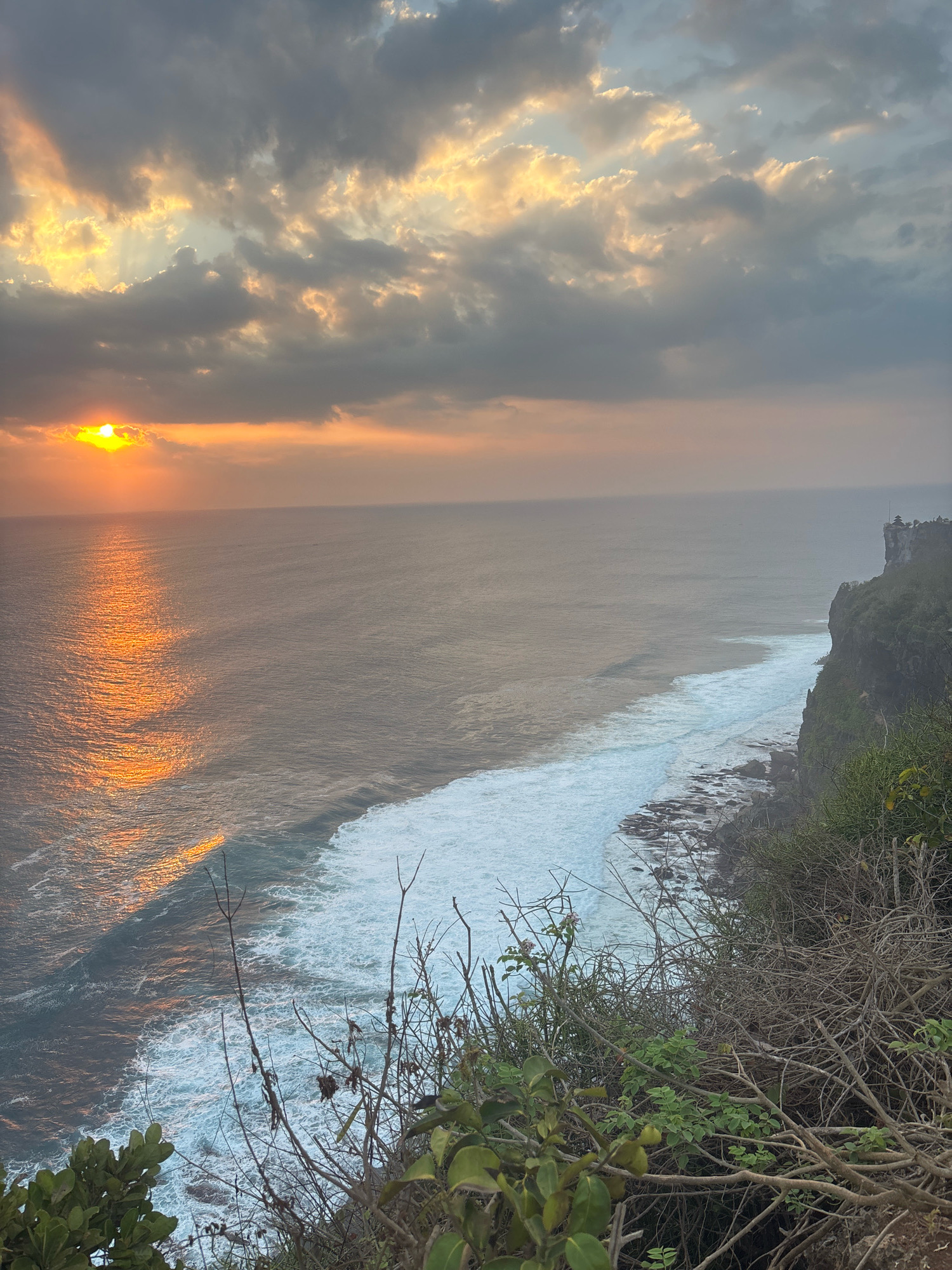 The image shows a cliffside view of the ocean at sunset, likely in Kuta Selatan, Indonesia. The sun, a bright orange orb, is low on the horizon, casting a shimmering golden reflection across the water. Dark, dramatic clouds are scattered across the sky, with rays of sunlight breaking through in some areas. The ocean below is a deep blue, transitioning to a foamy white where waves crash against the rocky shoreline and the base of the cliff. The cliff face is covered in lush green vegetation, with some dry, tangled brush in the foreground. A small temple or structure is visible atop the distant cliff. The overall impression is one of natural beauty and dramatic natural lighting.